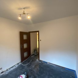 ANDRIY ANDRIISHYN - Pokój po odświeżeniu ścian na biało, zabezpieczona podłoga folią, widoczny otwarty, drewniany  portal drzwiowy i lampa sufitowa z dwoma reflektorami.