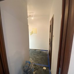 ANDRIY ANDRIISHYN - Korytarz po odświeżeniu: pomalowane ściany na biało, zabezpieczona podłoga folią, widoczny odkurzacz przemysłowy. Ujęcie z perspektywy otwartych drzwi.