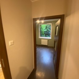 ANDRIY ANDRIISHYN - Korytarz z widokiem na pokój z oknem, przez które widać zieleń. Ściany w odcieniach beżu, podłoga ciemna. Widoczny fragment drzwi i włącznik światła.