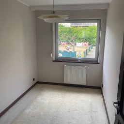 ANDRIY ANDRIISHYN - Jasny pokój z oknem na plac zabaw. Kremowa wykładzina, białe ściany, grzejnik pod oknem. Lampa z plisowanym abażurem. Widok na zieleń i bawiące się osoby.