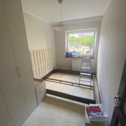 ANDRIY ANDRIISHYN - Rama łóżka z wezgłowiem w pokoju przed montażem, widoczna drabina, szafa wnękowa i okno z widokiem na zieleń. Neutralna kolorystyka wnętrza.