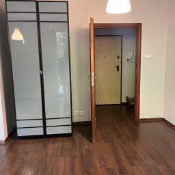 ANDRIY ANDRIISHYN - Pusty pokój po remoncie: brązowe panele, białe ściany, szafa z przesuwnymi drzwiami z mlecznego szkła i otwarte drzwi wejściowe. Widok na korytarz.