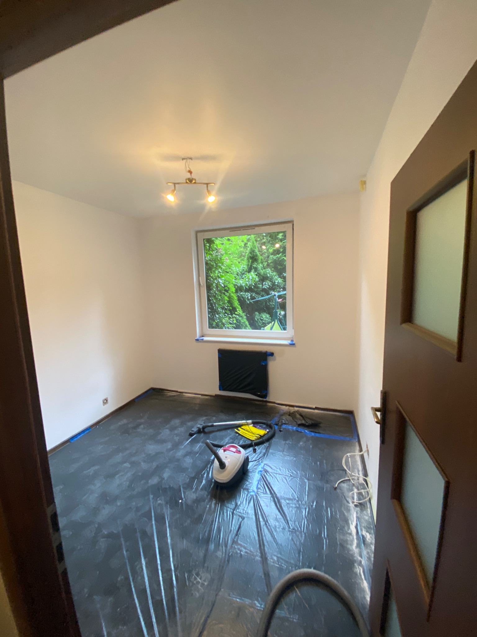 Pokój zabezpieczony folią malarską przed pracami. Widoczny odkurzacz, taśma malarska i osłonięty grzejnik. Przygotowanie do prac wykończeniowych.