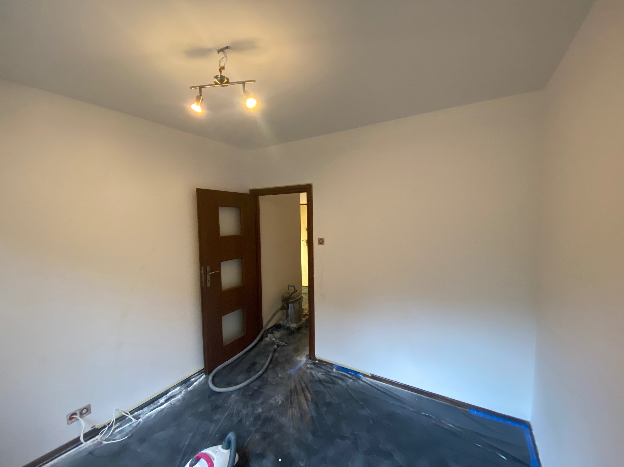 Pokój po odświeżeniu ścian na biało, zabezpieczona podłoga folią, widoczny otwarty, drewniany  portal drzwiowy i lampa sufitowa z dwoma reflektorami.