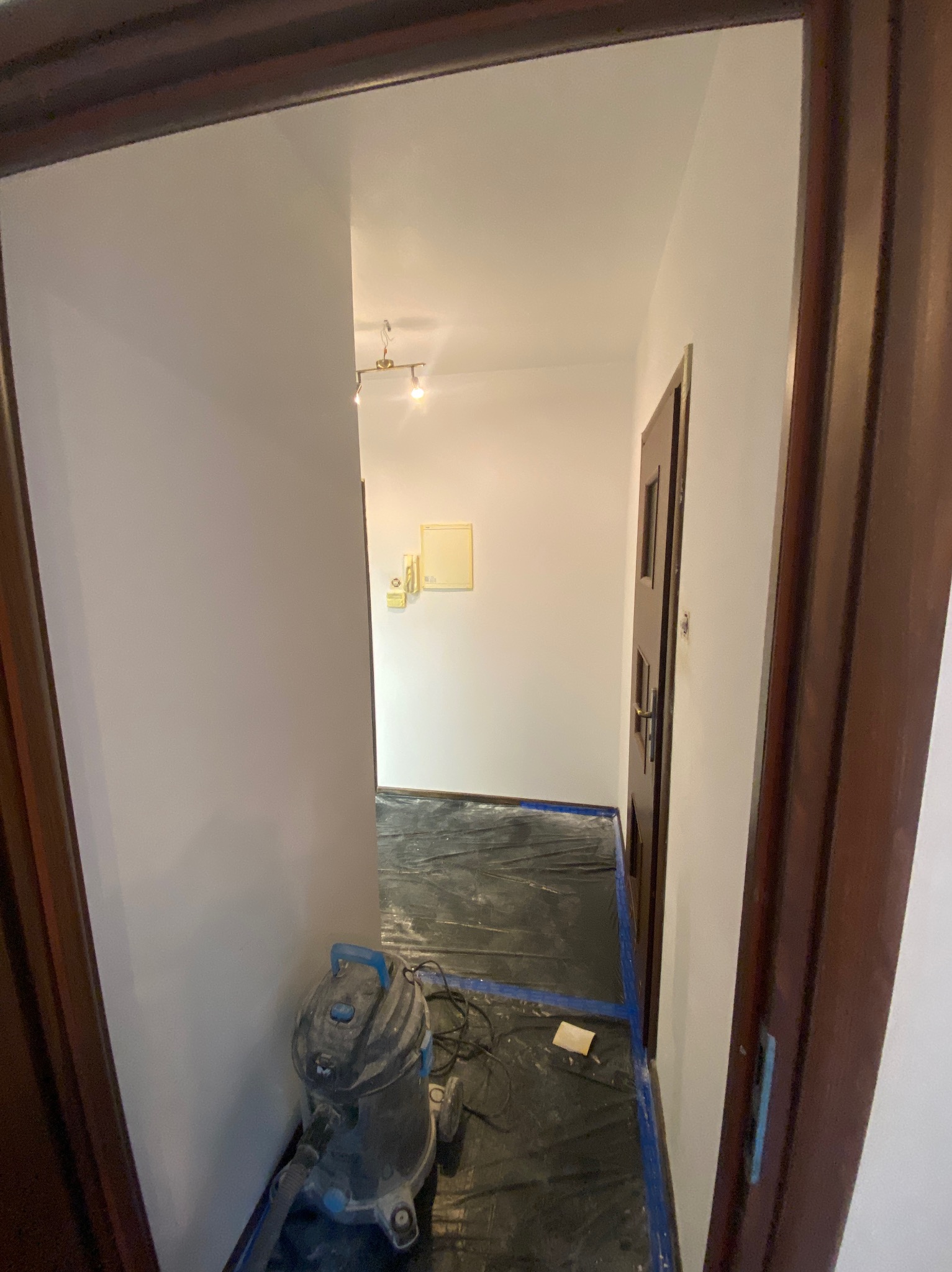 Korytarz po odświeżeniu: pomalowane ściany na biało, zabezpieczona podłoga folią, widoczny odkurzacz przemysłowy. Ujęcie z perspektywy otwartych drzwi.