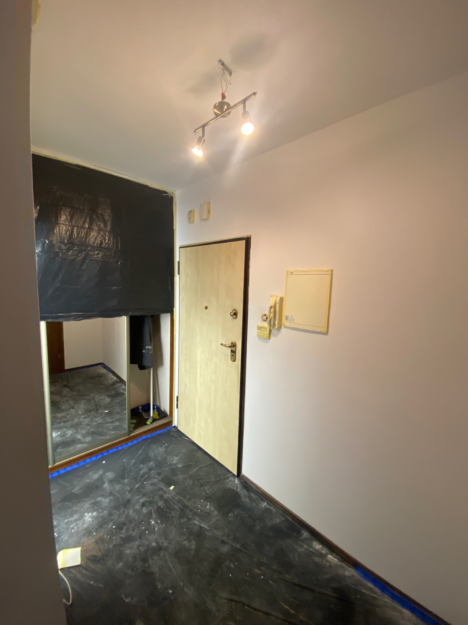 Korytarz w trakcie zabezpieczania folią malarską. Podłoga i framuga drzwi oklejone taśmą. Widoczna lampa sufitowa i domofon. Przygotowania do prac wykończeniowych.