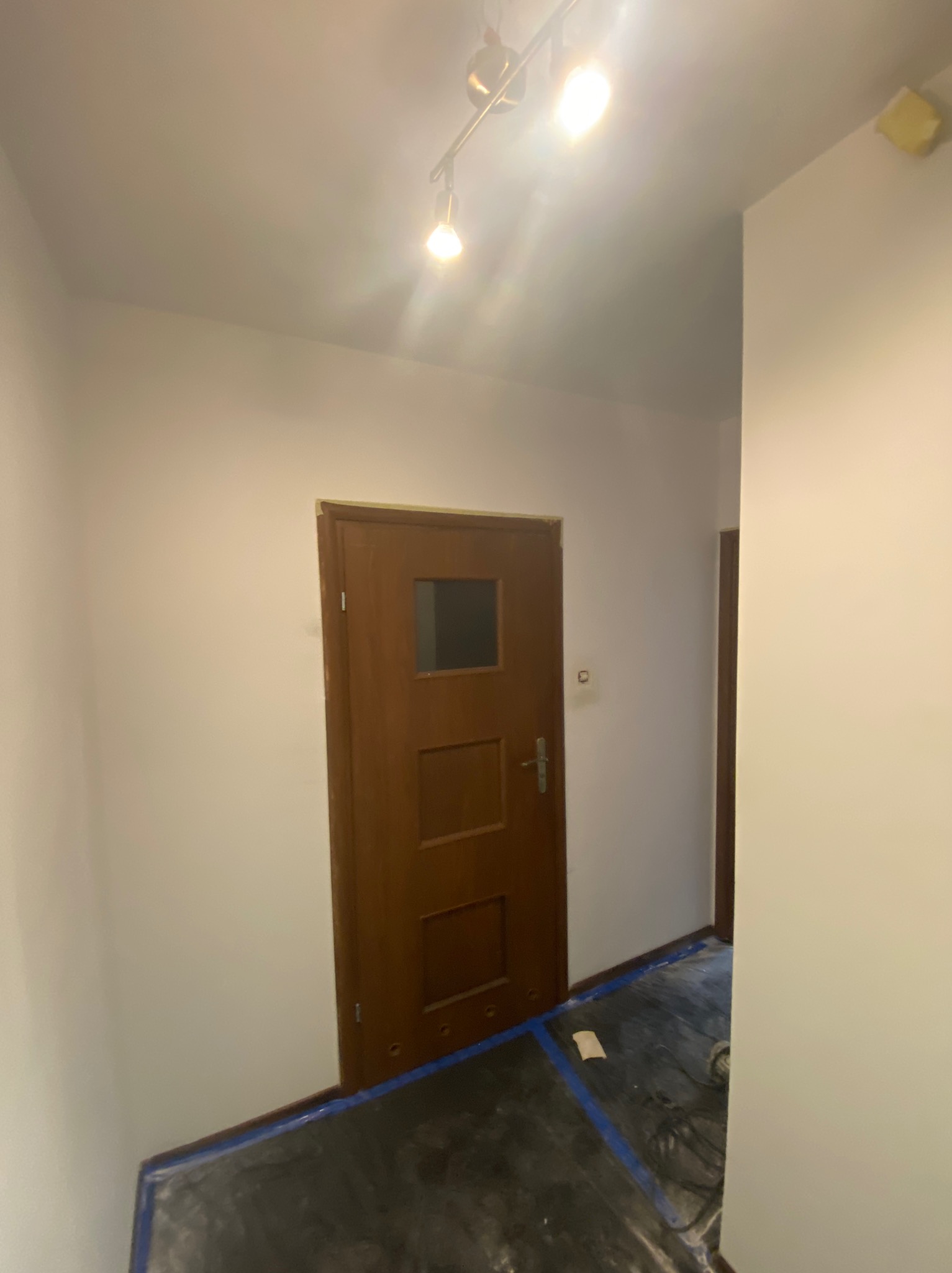 Świeżo pomalowane ściany w korytarzu, zabezpieczona podłoga folią malarską. Widoczne drewniane drzwi z szybą i lampa sufitowa.