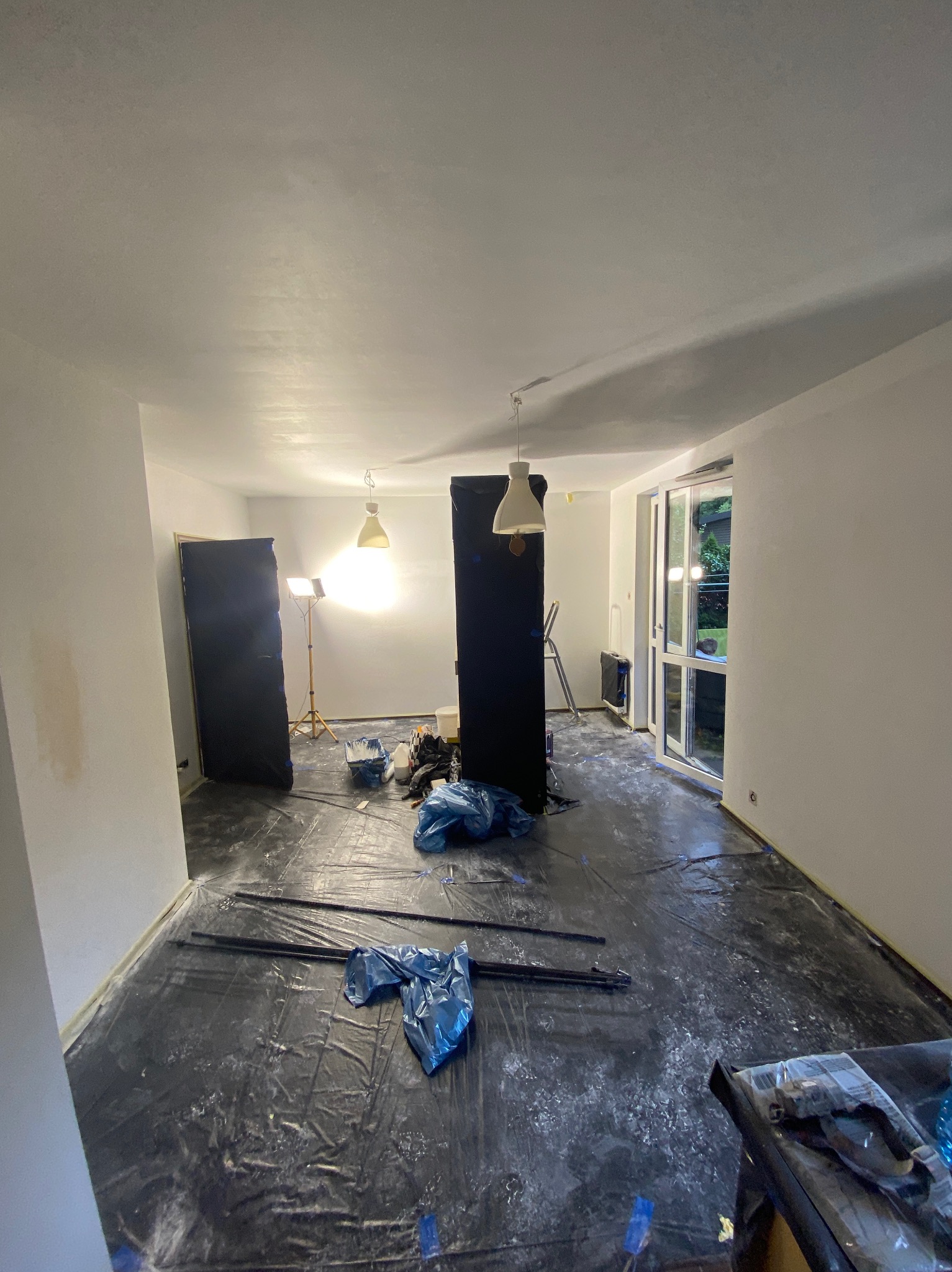 Pokój w trakcie przygotowań do malowania: podłoga zabezpieczona folią, widoczne narzędzia, lampa na statywie. Ściany świeżo pomalowane na biało.
