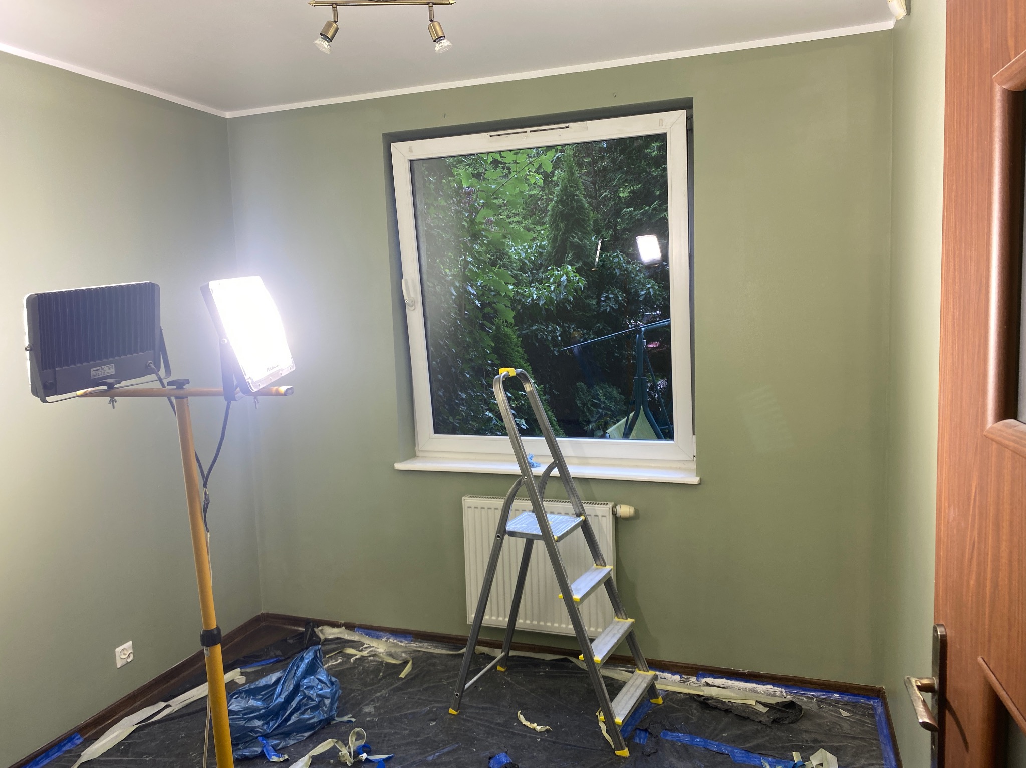 Ściana w odcieniach zieleni po malowaniu. Widoczna drabina, lampa budowlana i folia ochronna na podłodze. Okno z widokiem na zieleń.