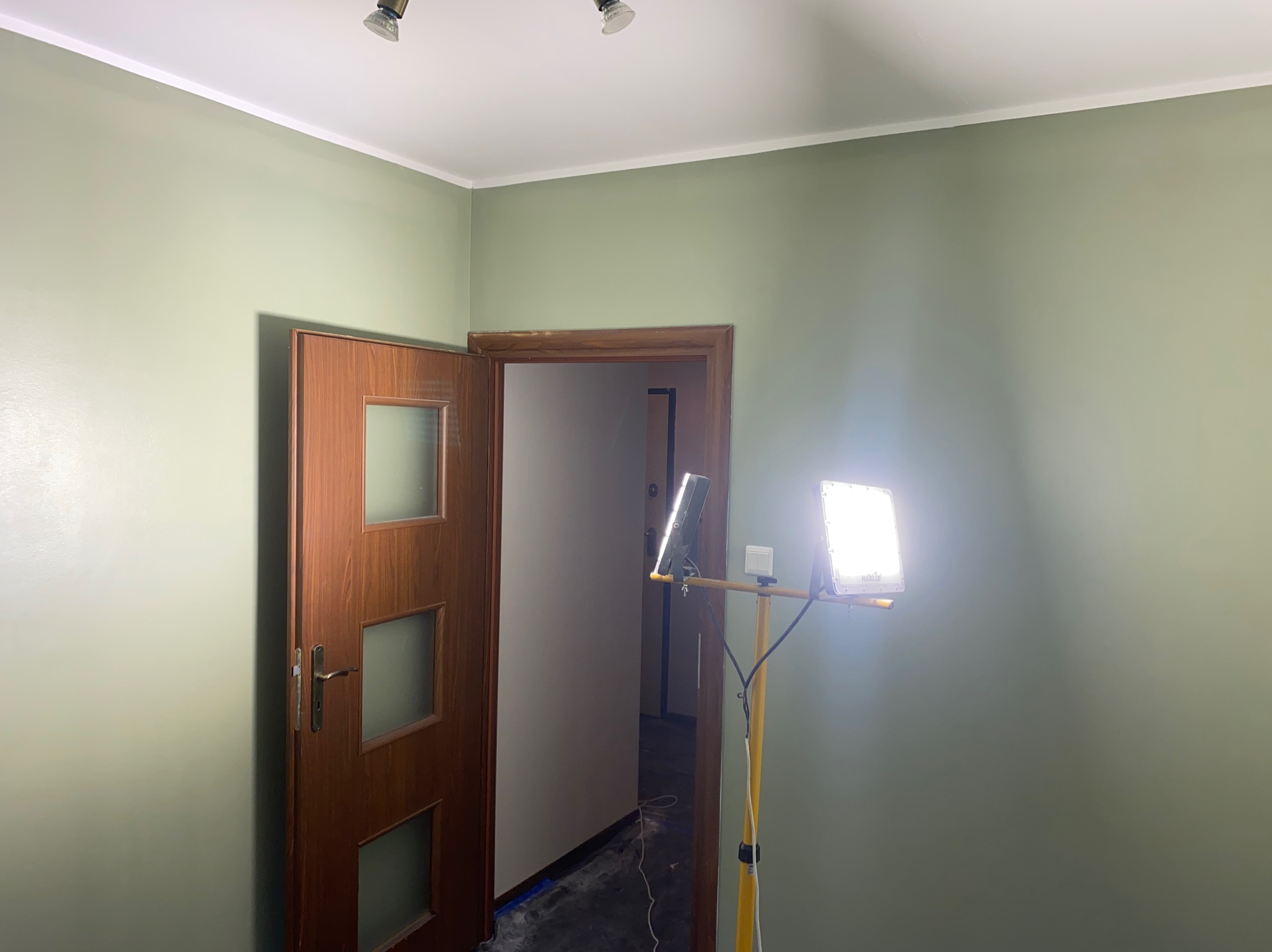 Pomalowane na zielono wnętrze z otwartymi drewnianymi drzwiami i lampą budowlaną na statywie. Widoczne listwy przysufitowe i dwa reflektory sufitowe.