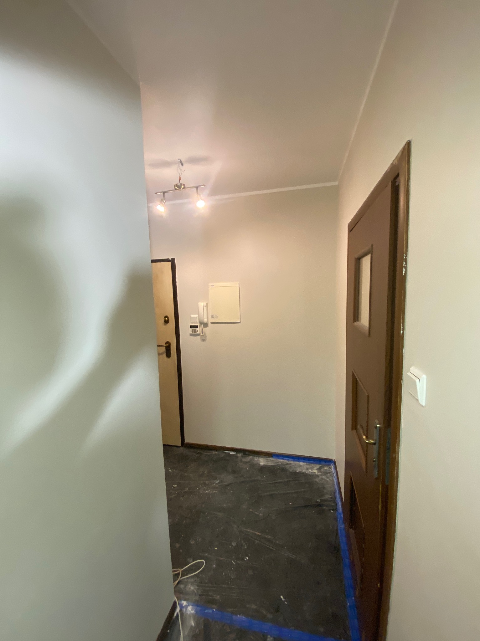 Korytarz w trakcie zabezpieczania folią malarską przed pracami. Widoczne drzwi wejściowe i wewnętrzne, ściany przygotowane do malowania.