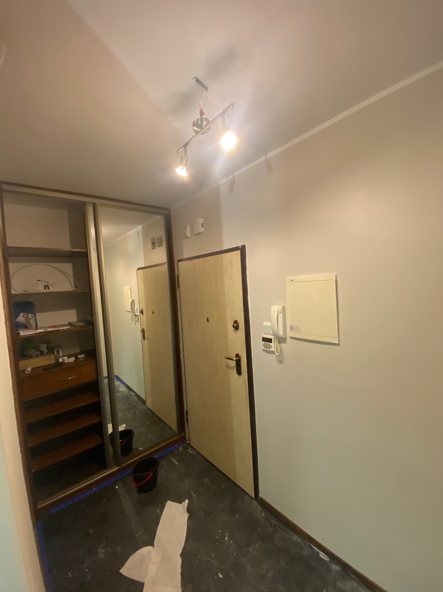 Korytarz po malowaniu: szara ściana, drzwi wejściowe, szafa z lustrem. Widoczne ślady farby na podłodze, taśma malarska i wiadro, co sugeruje świeżo zakończone prace.