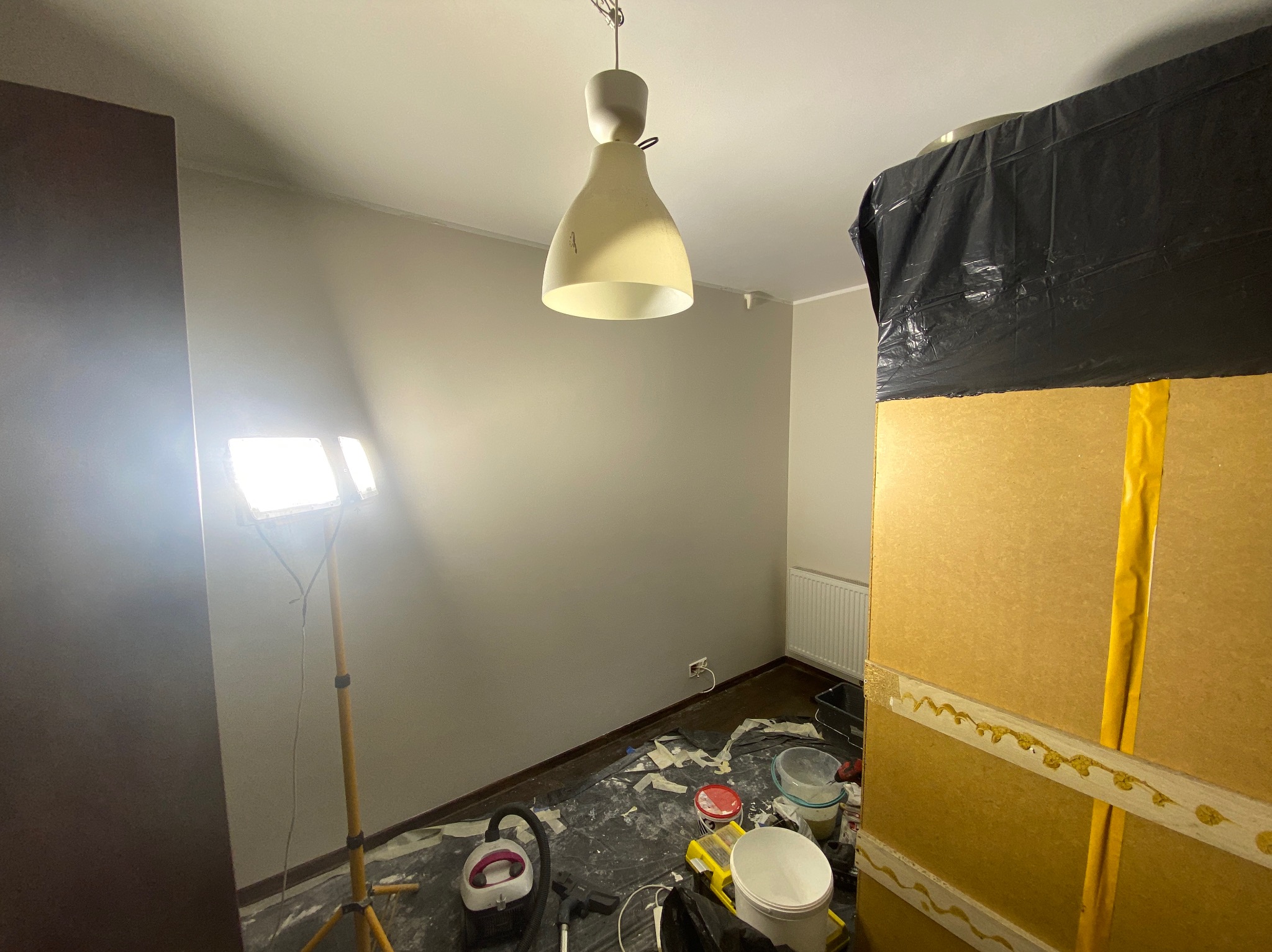 Pokój przygotowany do odświeżenia: zabezpieczone meble, rozłożona folia, pędzle, wałki i lampa na statywie. Ściany pomalowane na szaro.