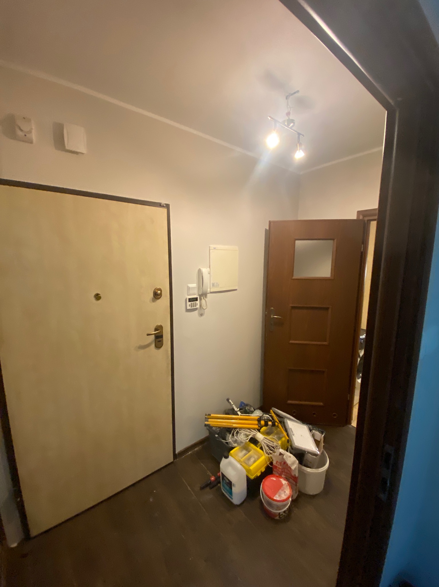 Korytarz w trakcie prac: widoczne drzwi wejściowe, drzwi wewnętrzne oraz narzędzia i materiały budowlane. Przy ścianie leżą pędzle, wiadra i inne akcesoria.