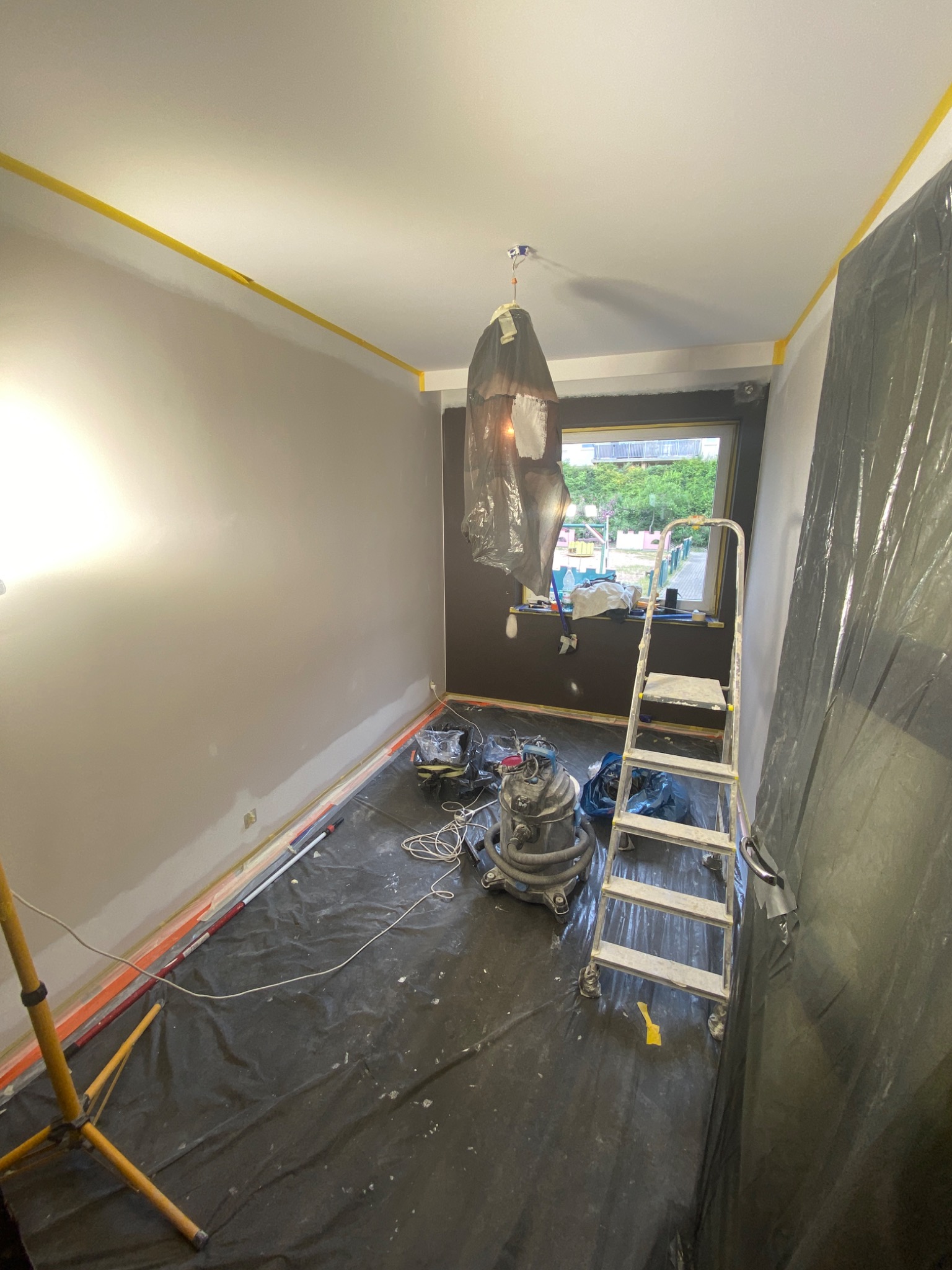 Pokój w trakcie odświeżania: ściany częściowo pomalowane na szaro, zabezpieczona folią podłoga, drabina i lampa pod sufitem osłonięta folią. Przygotowania do malowania.