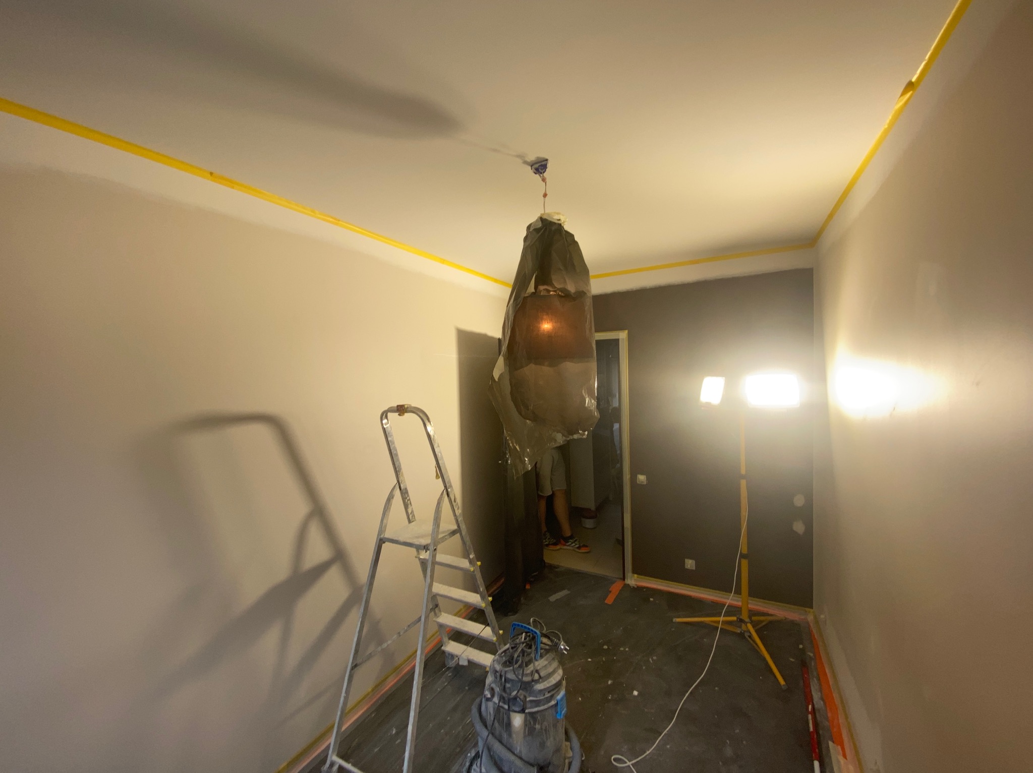 Pokój w trakcie prac: zabezpieczona lampa, drabina, odkurzacz, taśma malarska na styku ściany i sufitu, ściany częściowo pomalowane na szaro.