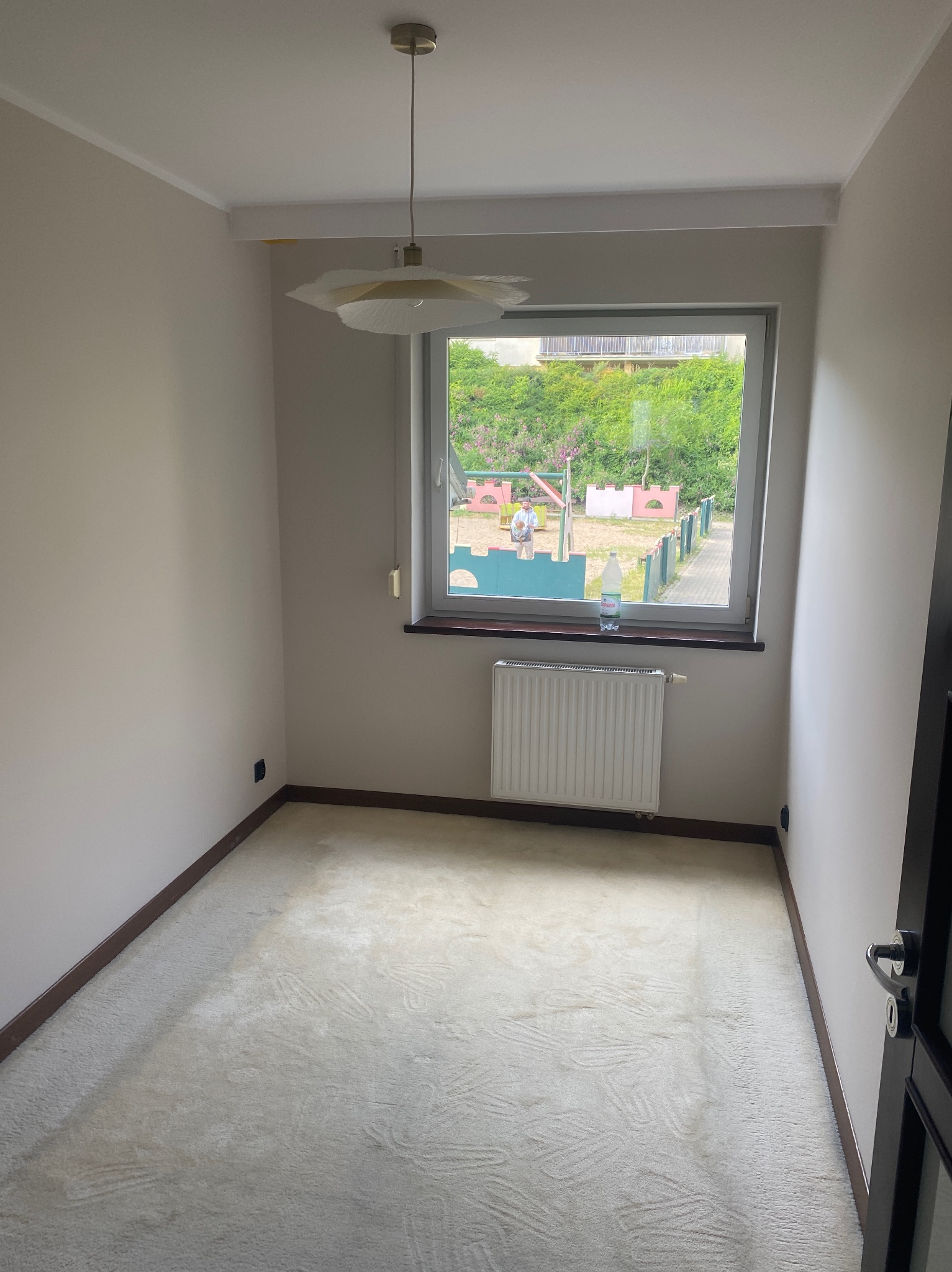 Jasny pokój z oknem na plac zabaw. Kremowa wykładzina, białe ściany, grzejnik pod oknem. Lampa z plisowanym abażurem. Widok na zieleń i bawiące się osoby.