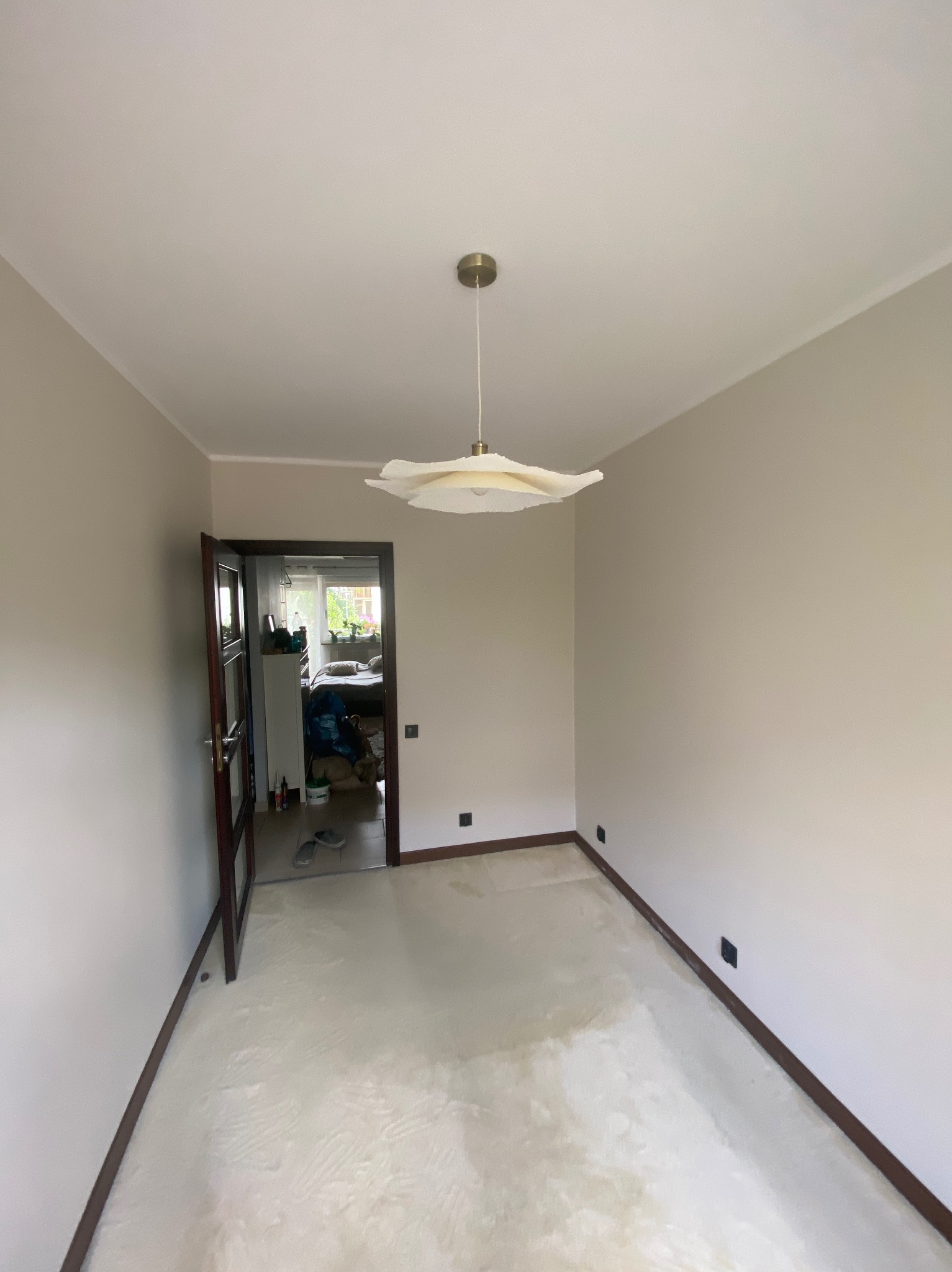 Jasne, świeżo pomalowane wnętrze z otwartymi drzwiami, przez które widać fragment pokoju. W centralnej części wisi nowoczesna, biała lampa.