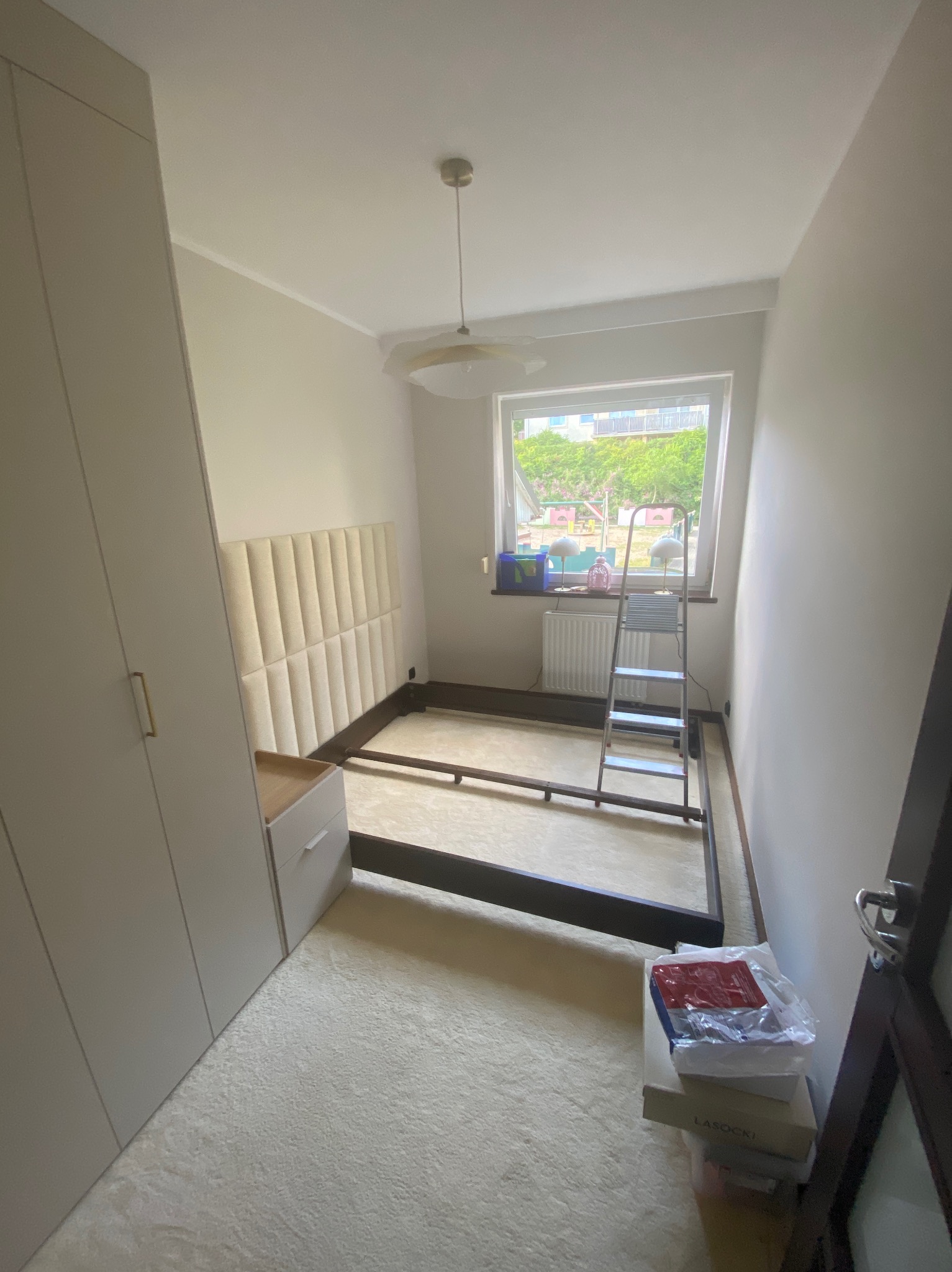 Rama łóżka z wezgłowiem w pokoju przed montażem, widoczna drabina, szafa wnękowa i okno z widokiem na zieleń. Neutralna kolorystyka wnętrza.