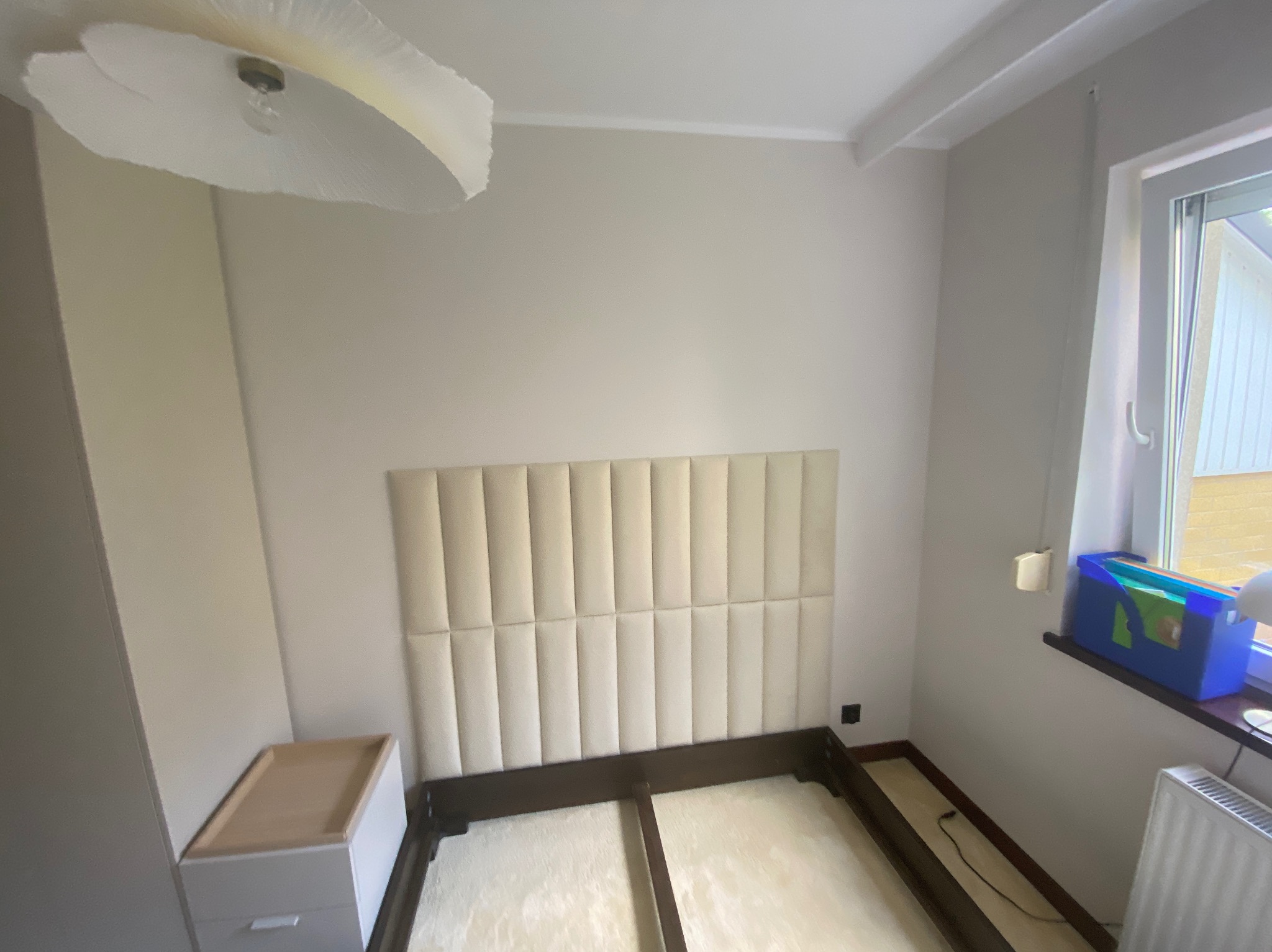 Sypialnia z beżowym tapicerowanym zagłówkiem łóżka i minimalistyczną białą szafką nocną, oświetlona lampą sufitową w kształcie kwiatu.