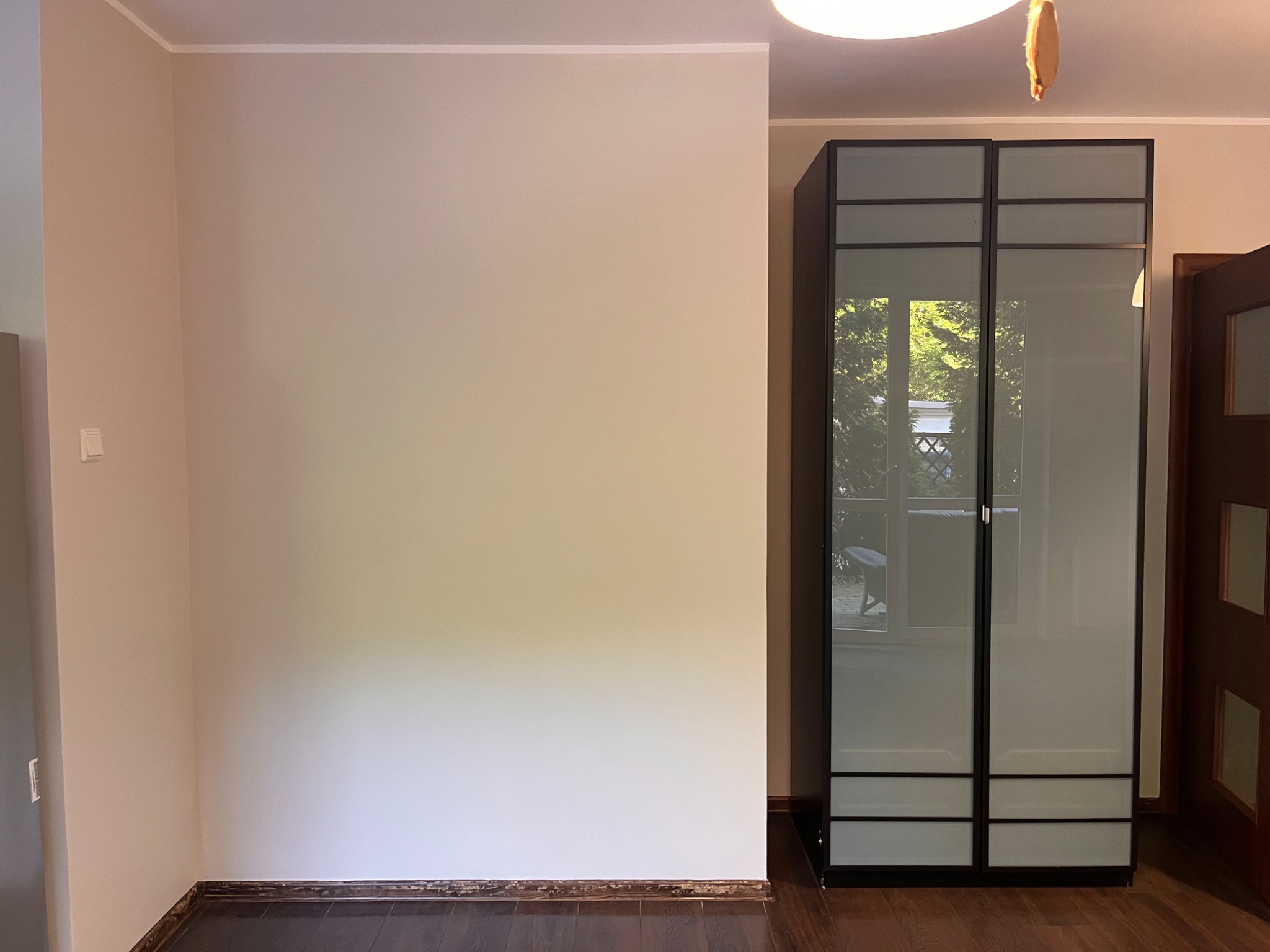 Jasny pokój po odświeżeniu ścian z drewnianą podłogą, z szafą z przesuwnymi drzwiami z mlecznego szkła i czarnymi ramami. Widok na zewnątrz przez odbicie w szkle.