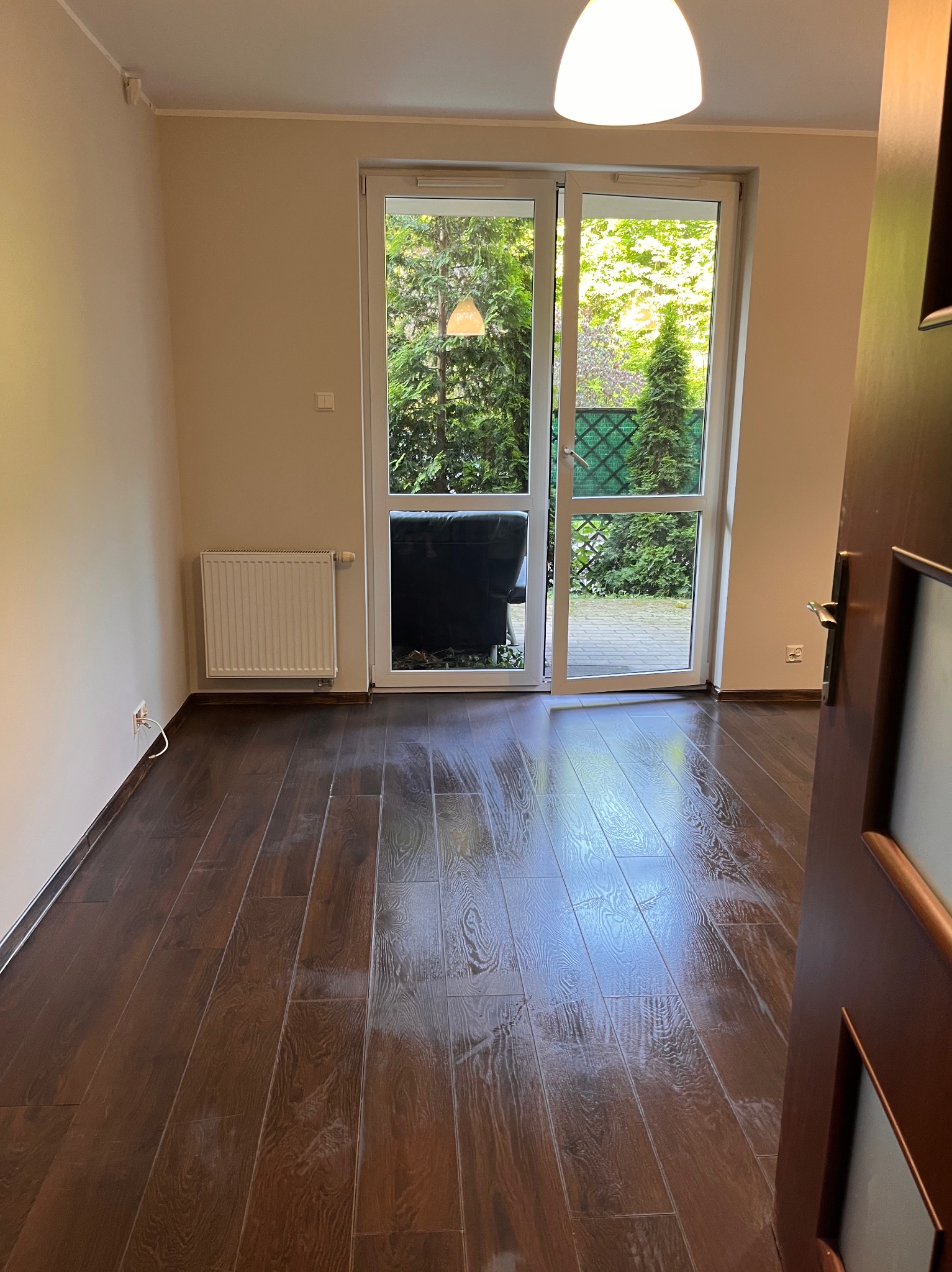 Jasny pokój z ciemną, błyszczącą podłogą, białymi ścianami i grzejnikiem. Widok na zieleń przez przeszklone drzwi balkonowe. Częściowo otwarte drzwi po prawej.