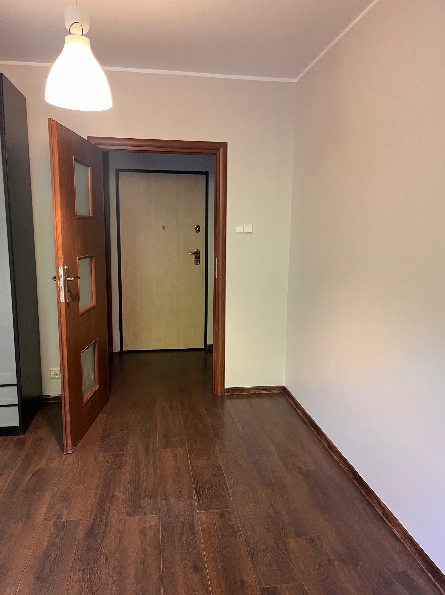 Pusty pokój po remoncie: ciemne panele podłogowe, jasne ściany i otwarte drewniane drzwi z szybkami, widok na kolejne drzwi. Lampa sufitowa w rogu.