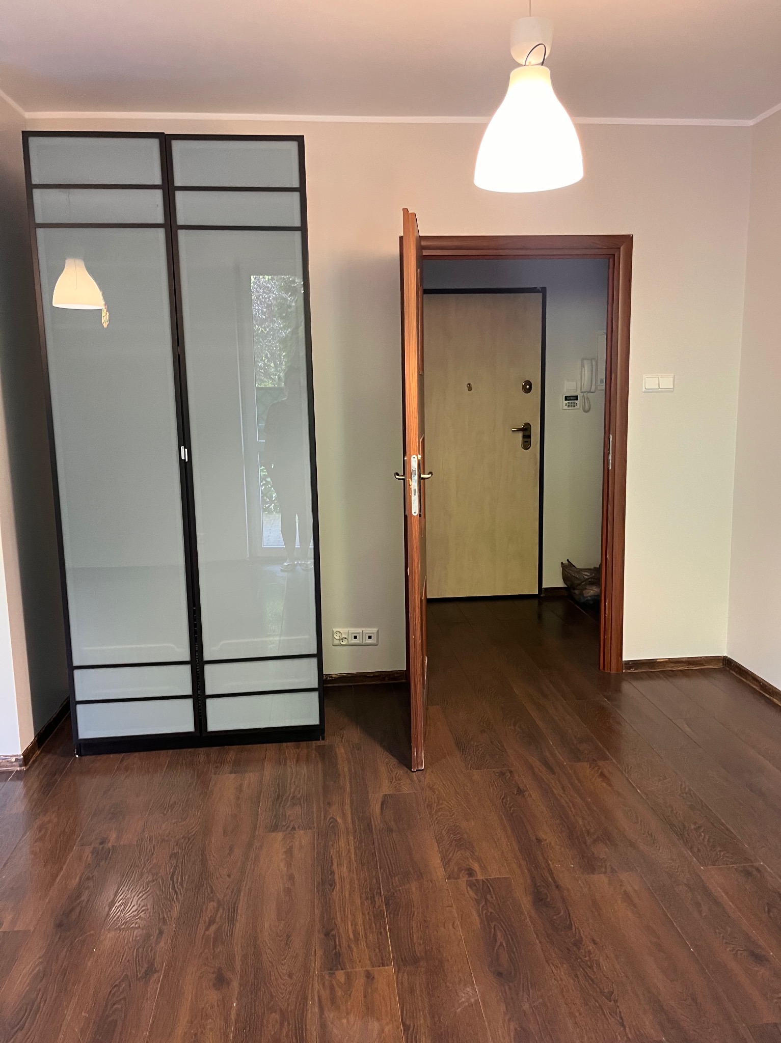 Pusty pokój po remoncie: brązowe panele, białe ściany, szafa z przesuwnymi drzwiami z mlecznego szkła i otwarte drzwi wejściowe. Widok na korytarz.