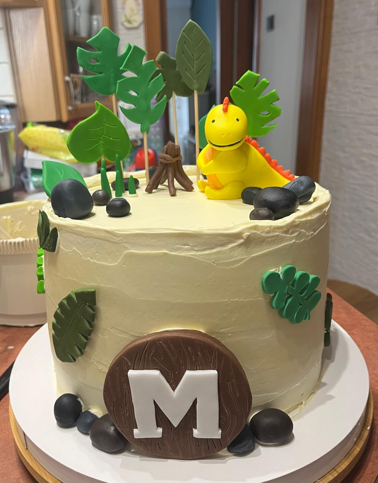 Tort urodzinowy z motywem dinozaura i inicjałem 'M', ozdobiony liśćmi z lukru, czarnymi kamykami i ogniskiem, ustawiony na białym talerzu.