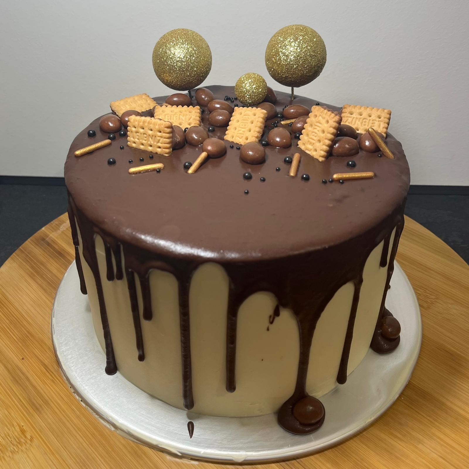 Tort z czekoladową polewą, dekorowany biszkoptami, czekoladowymi drażami i trzema złotymi kulami na patyczkach, umieszczony na drewnianej desce.