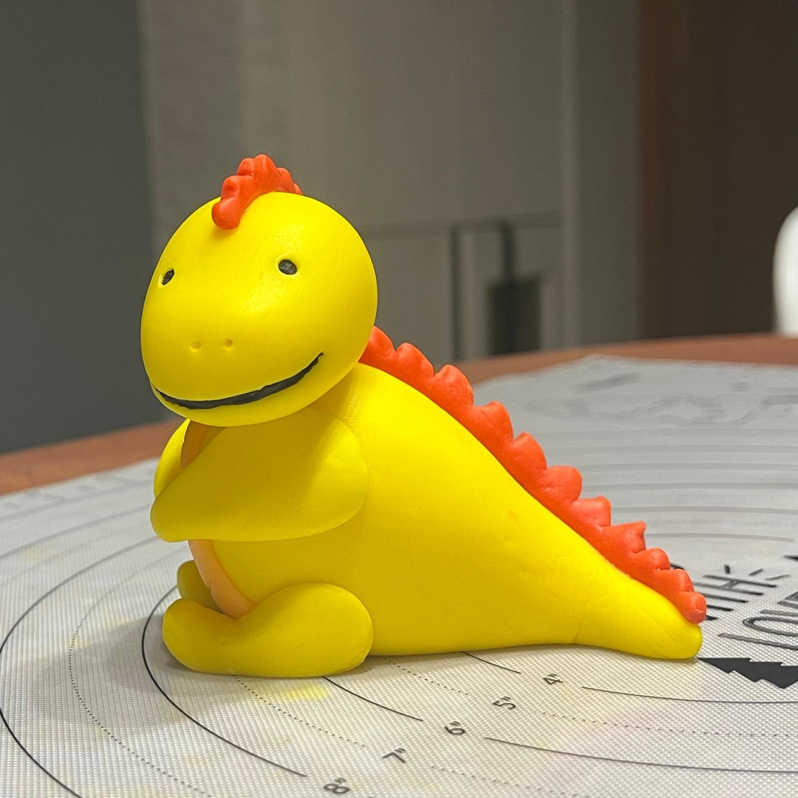 Żółty dinozaur z czerwonymi kolcami wykonany z masy cukrowej, siedzący na macie silikonowej z podziałką.