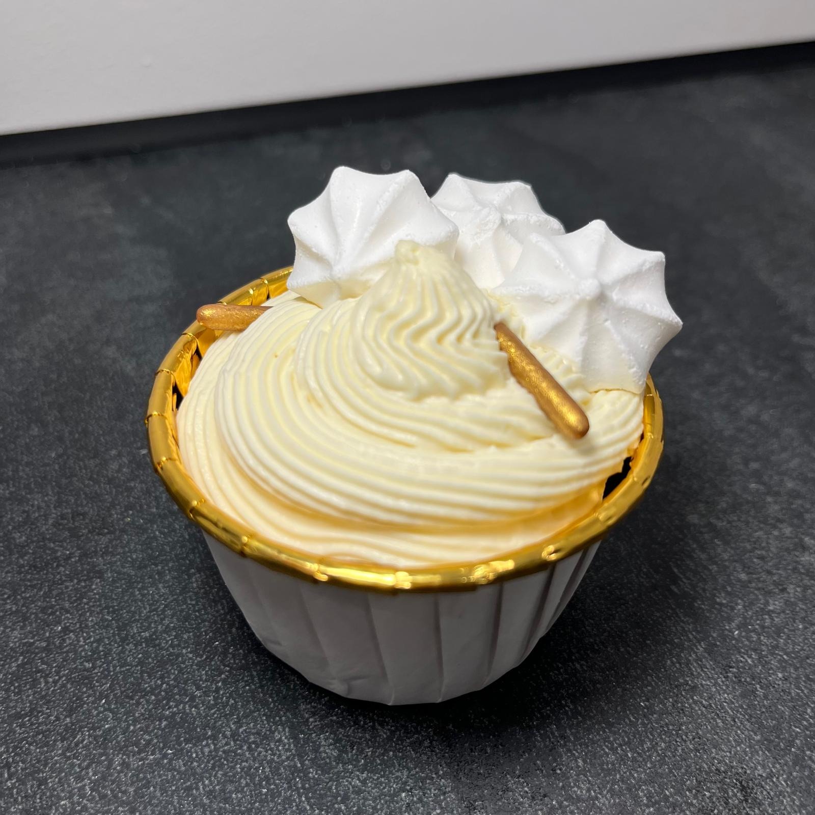 Kremowy babeczka z białymi bezami i złotymi posypkami, umieszczona w papilotce z złotym obramowaniem, na ciemnym blacie.