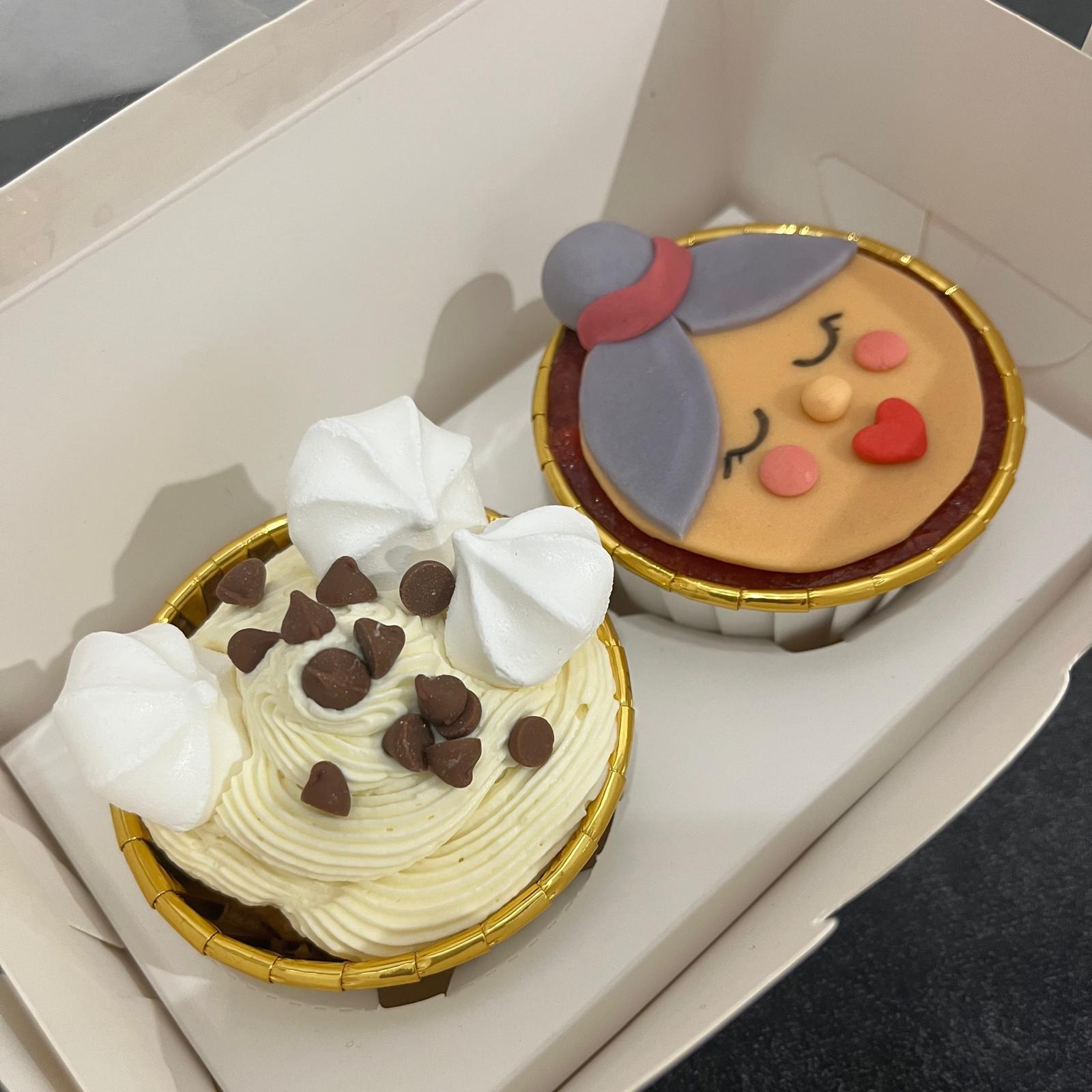 Dwa ozdobne babeczki w papierowych foremkach z kremem i dekoracjami, w białym pudełku: jeden z bitą śmietaną, czekoladowymi groszkami i bezami, drugi z lukrowaną postacią śpiącej starszej pani...