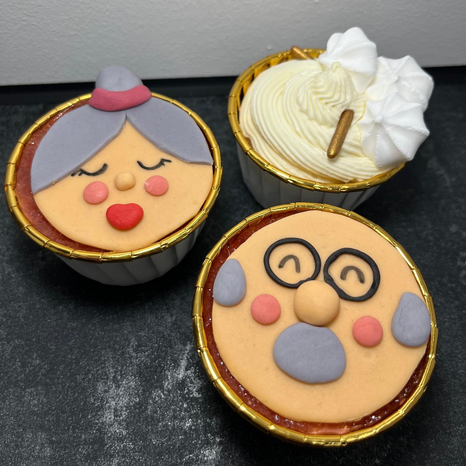 Trzy babeczki w papilotkach z dekoracjami z masy cukrowej: dwie z postaciami starszych osób (kobieta w czepku i mężczyzna w okularach), jedna z kremem i bezami.