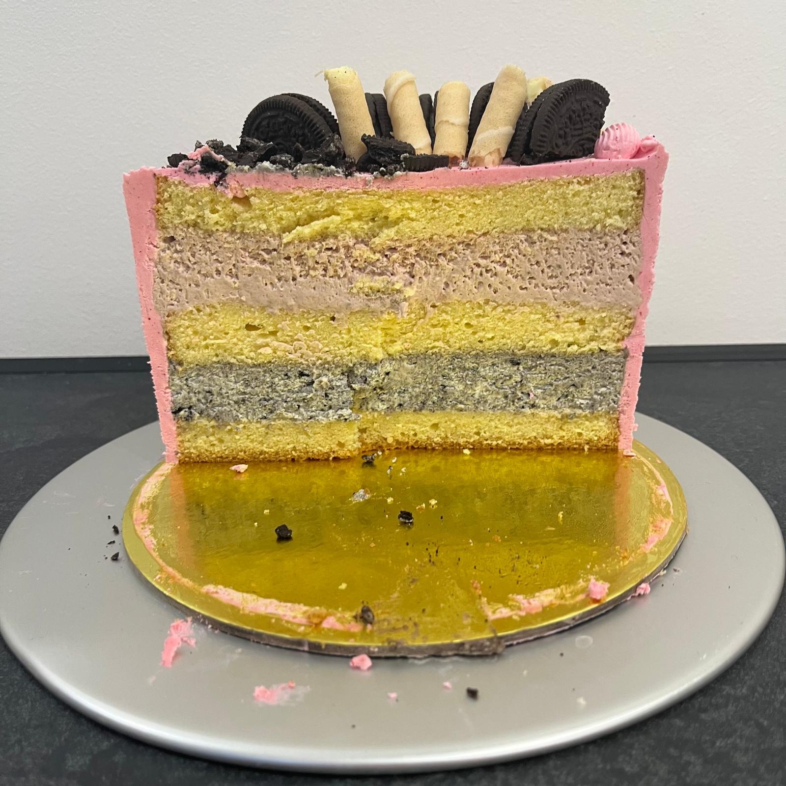Przekrój tortu z różowym kremem, warstwami biszkoptu w odcieniach żółci i szarości, udekorowanego Oreo i waflami na złotej tacy.