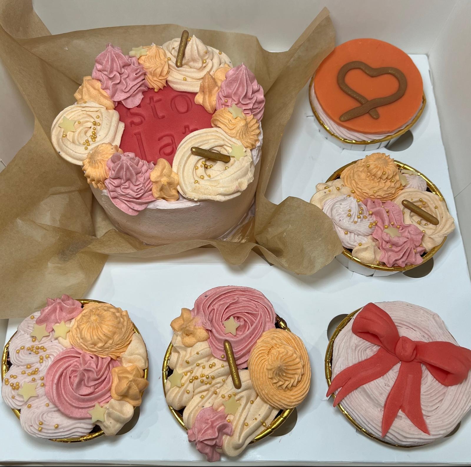 Pudełko z tortem urodzinowym i czterema babeczkami, dekorowane kremem w odcieniach różu i pomarańczy, posypane złotym brokatem, z elementami w kształcie gwiazdek i serca.