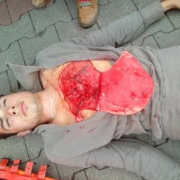 MISJA PP Daria Jasnoch-Klupś - Młody mężczyzna w szarej bluzie leżący na chodniku z symulowaną raną klatki piersiowej, obok widoczny fragment pomarańczowych noszy i czyjeś buty.