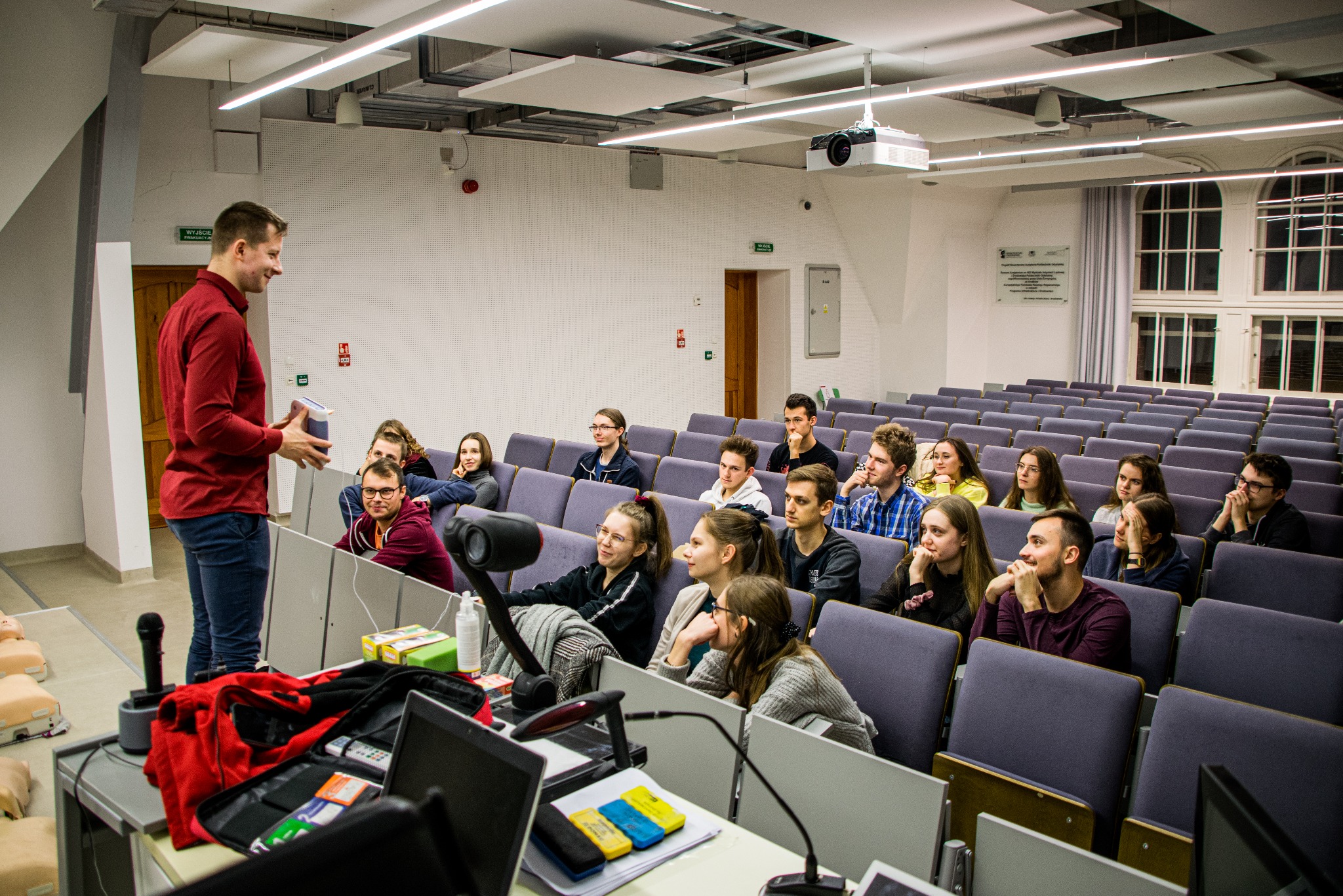 Instruktor w czerwonej koszuli prowadzi szkolenie z zakresu ratownictwa medycznego dla grupy osób siedzących w sali wykładowej w Gdyni. Na pierwszym planie widoczne manekiny do ćwiczeń resuscytacji...