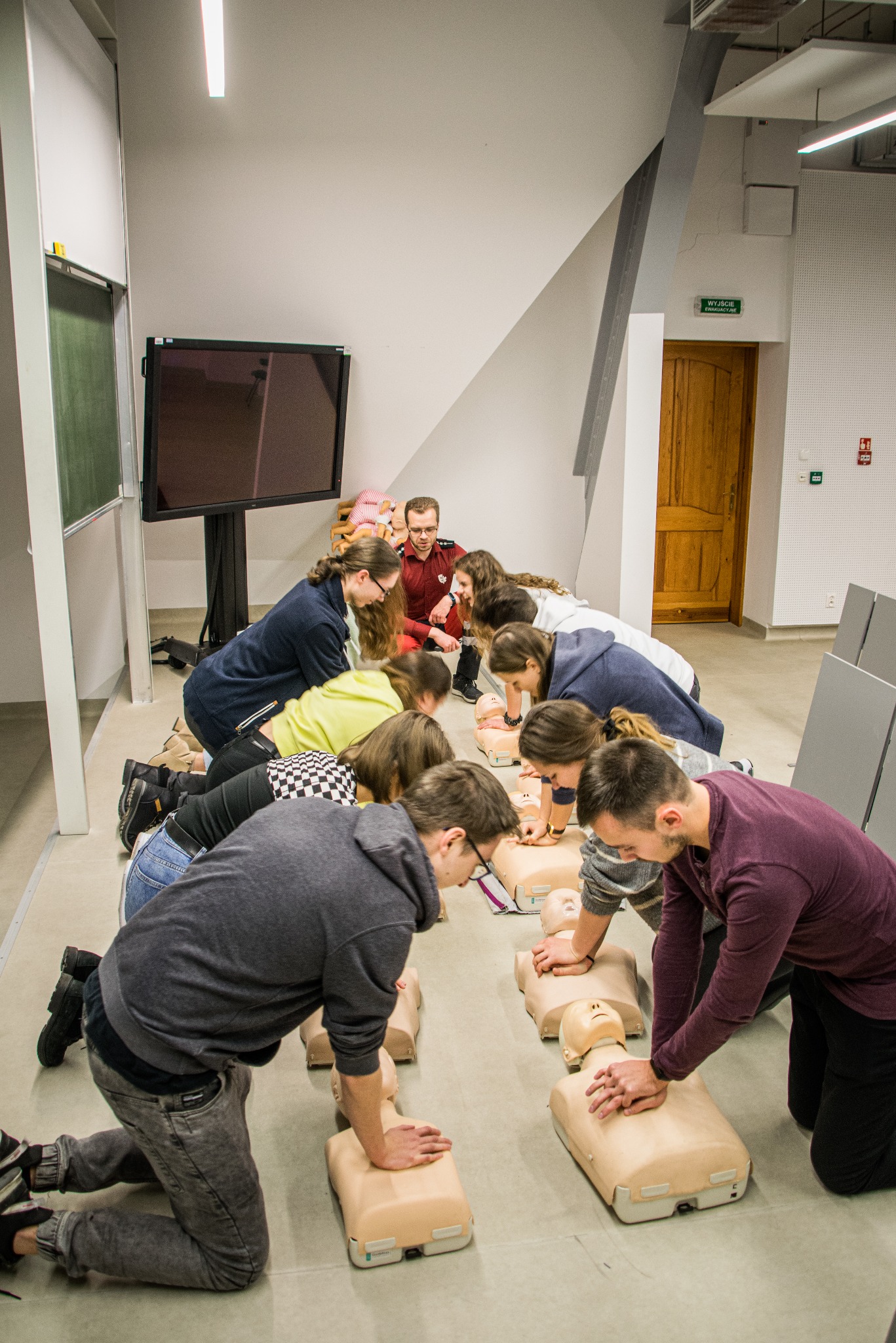 Grupa osób ćwiczy resuscytację krążeniowo-oddechową na manekinach pod nadzorem instruktora w czerwonej koszuli, w tle tablica interaktywna i drzwi ewakuacyjne.
