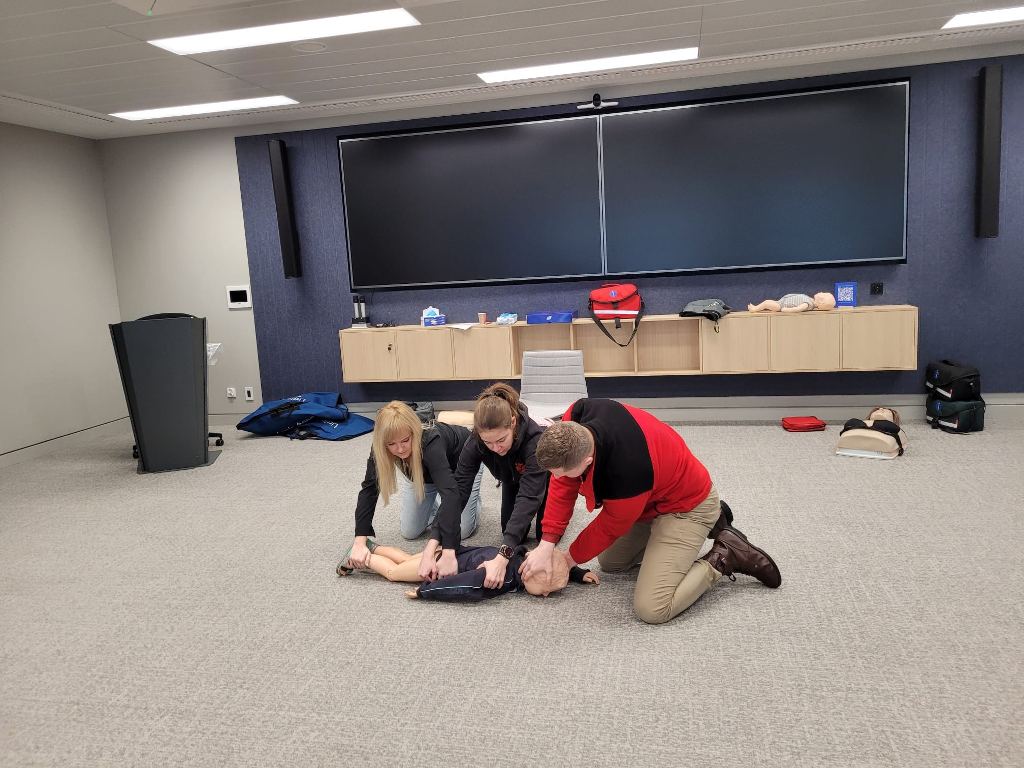 Trzy osoby ćwiczą resuscytację krążeniowo-oddechową na manekinie niemowlęcia w sali szkoleniowej z dużym ekranem i sprzętem medycznym w tle.