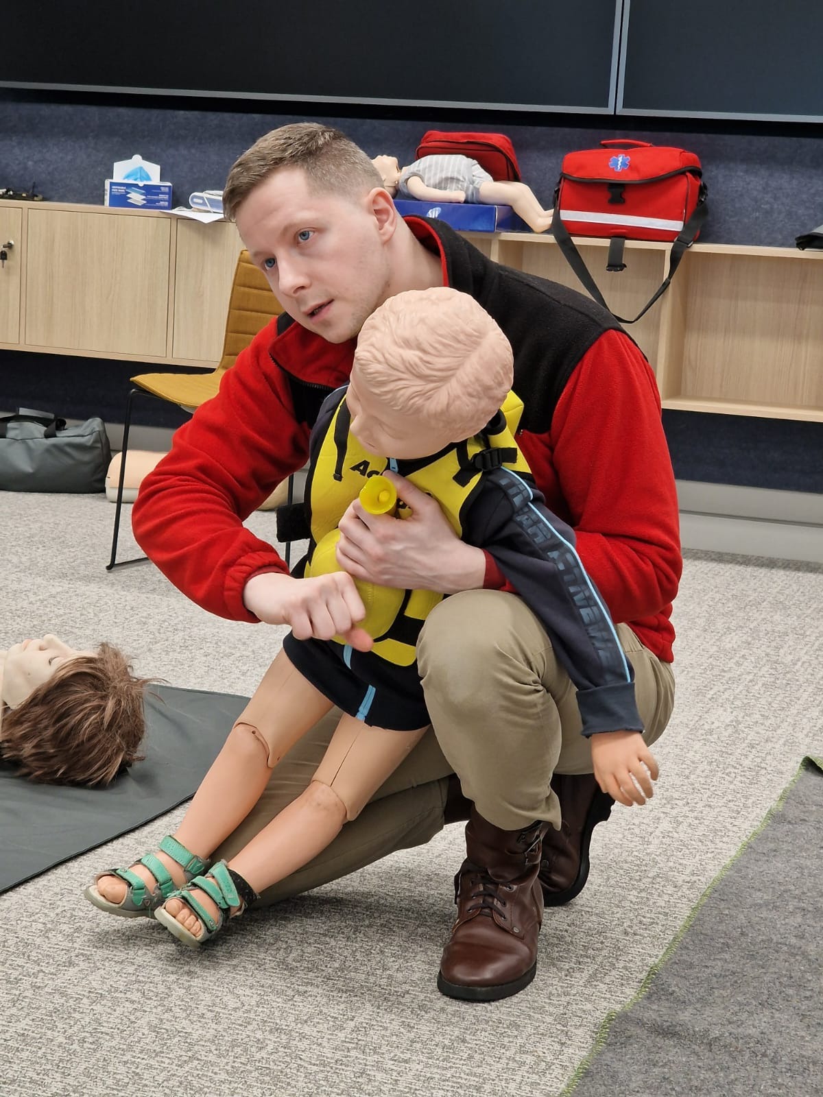 Instruktor w czerwonej bluzie demonstruje resuscytację krążeniowo-oddechową na manekinie dziecka ubranego w żółtą kamizelkę, w tle widoczna apteczka i inne manekiny.