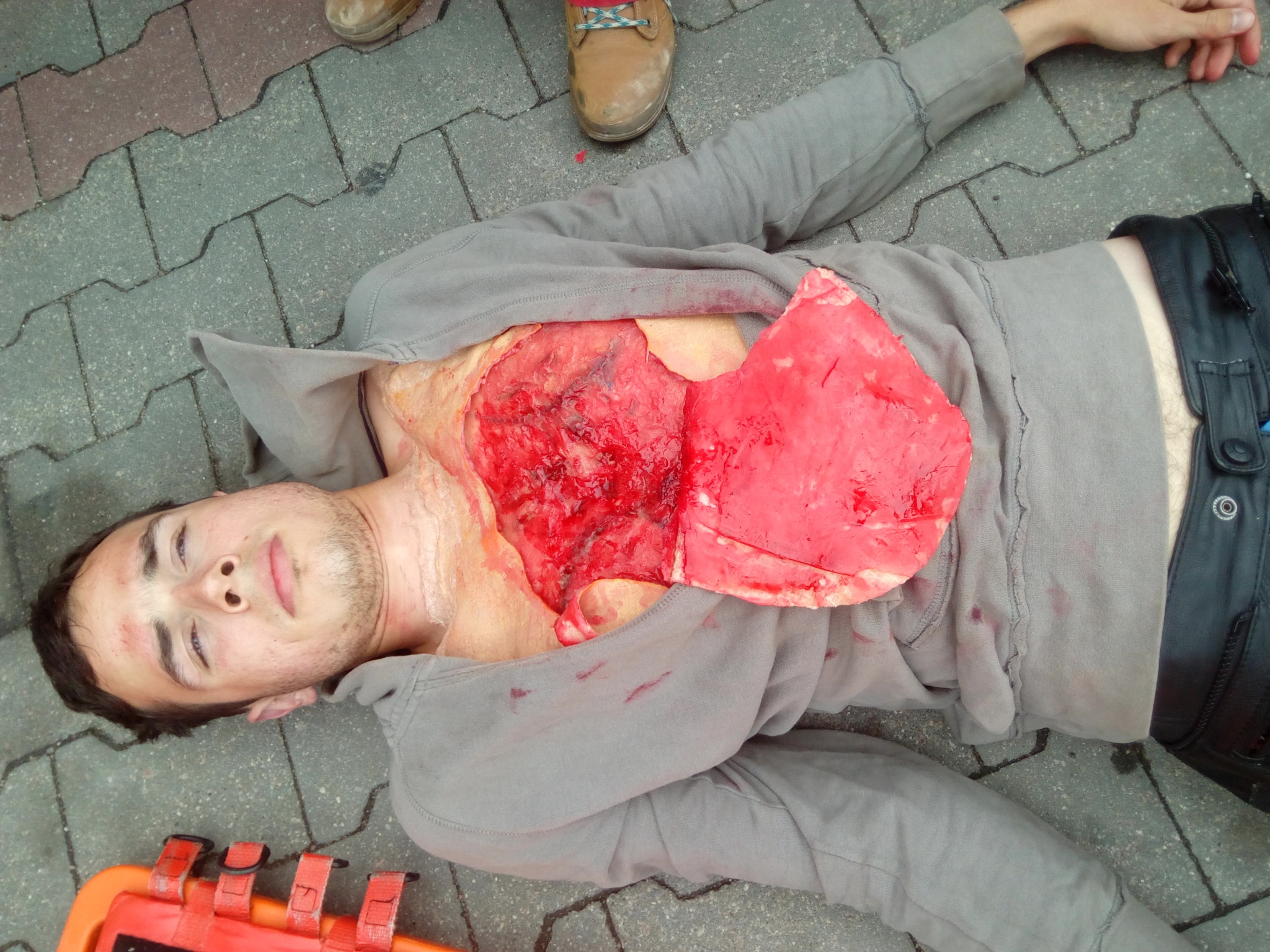 Młody mężczyzna w szarej bluzie leżący na chodniku z symulowaną raną klatki piersiowej, obok widoczny fragment pomarańczowych noszy i czyjeś buty.