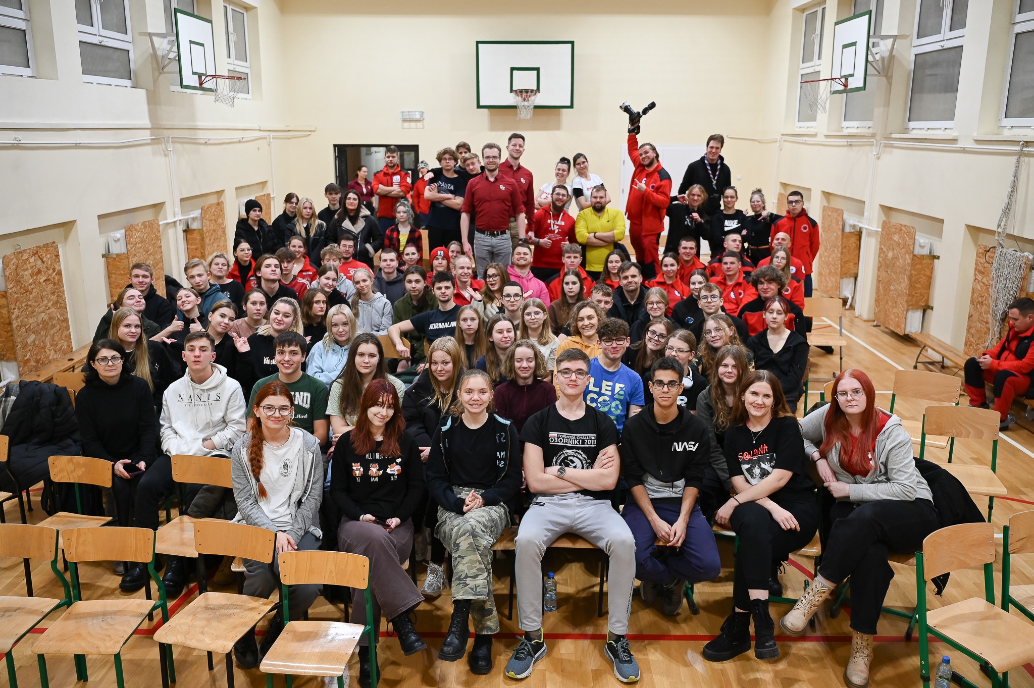 Duża grupa osób, uczestników kursu pierwszej pomocy i instruktorów, pozuje do grupowego zdjęcia w sali gimnastycznej z koszem do koszykówki w tle.