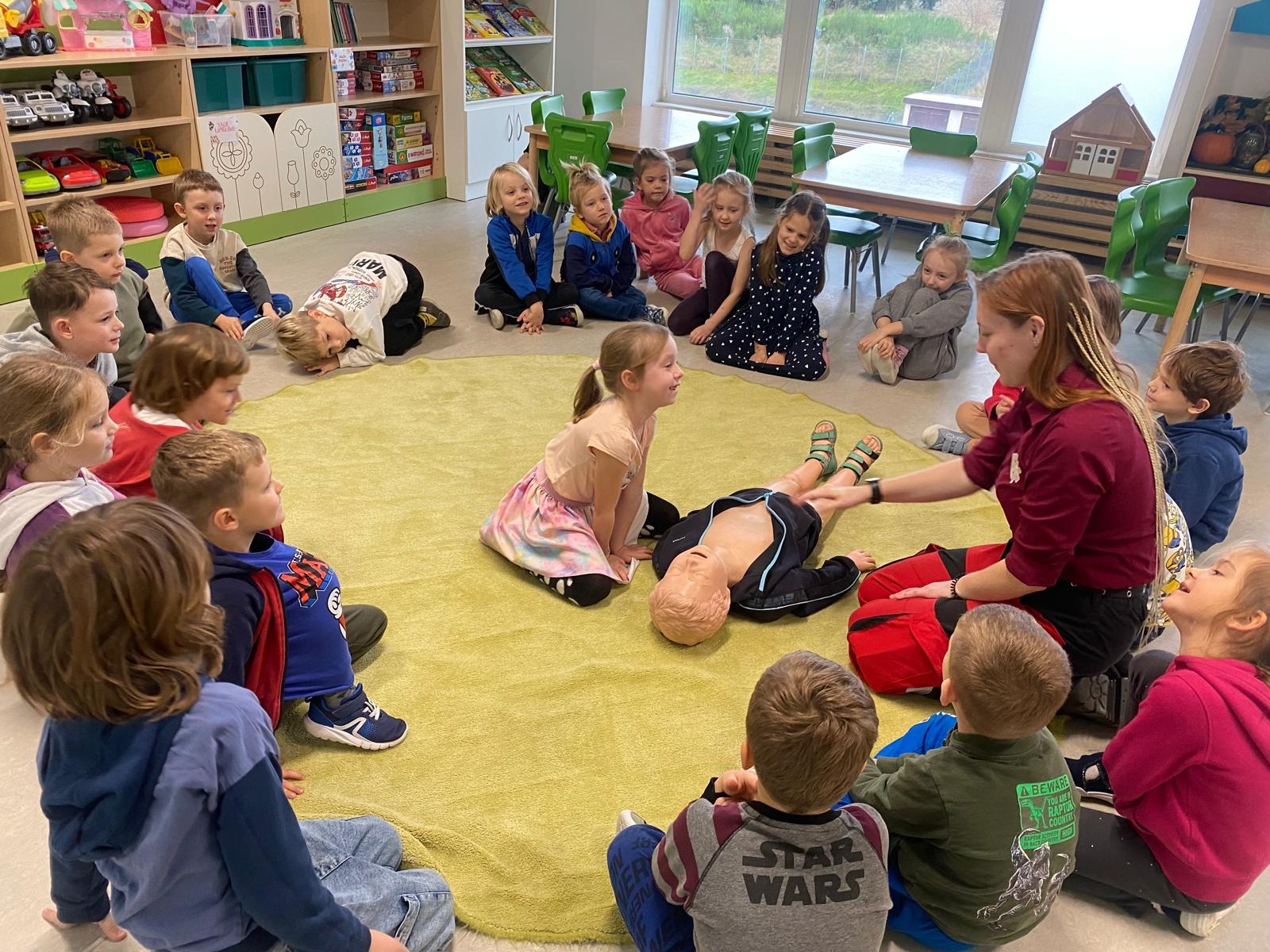 Instruktorka z blond warkoczykami demonstruje na manekinie resuscytację krążeniowo-oddechową, otoczona grupą dzieci siedzących na okrągłym, jasnożółtym dywanie w sali przedszkolnej z regałami...