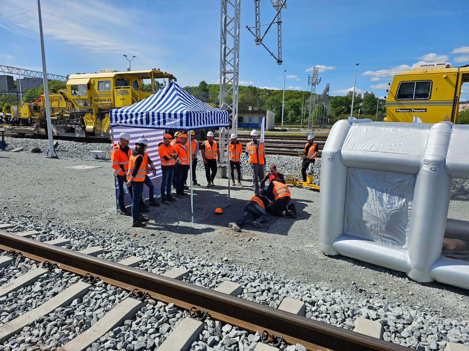 Symulacja akcji ratunkowej na terenie kolejowym: grupa osób w pomarańczowych kamizelkach udziela pomocy poszkodowanemu leżącemu na ziemi, w tle maszyny budowlane i namiot.