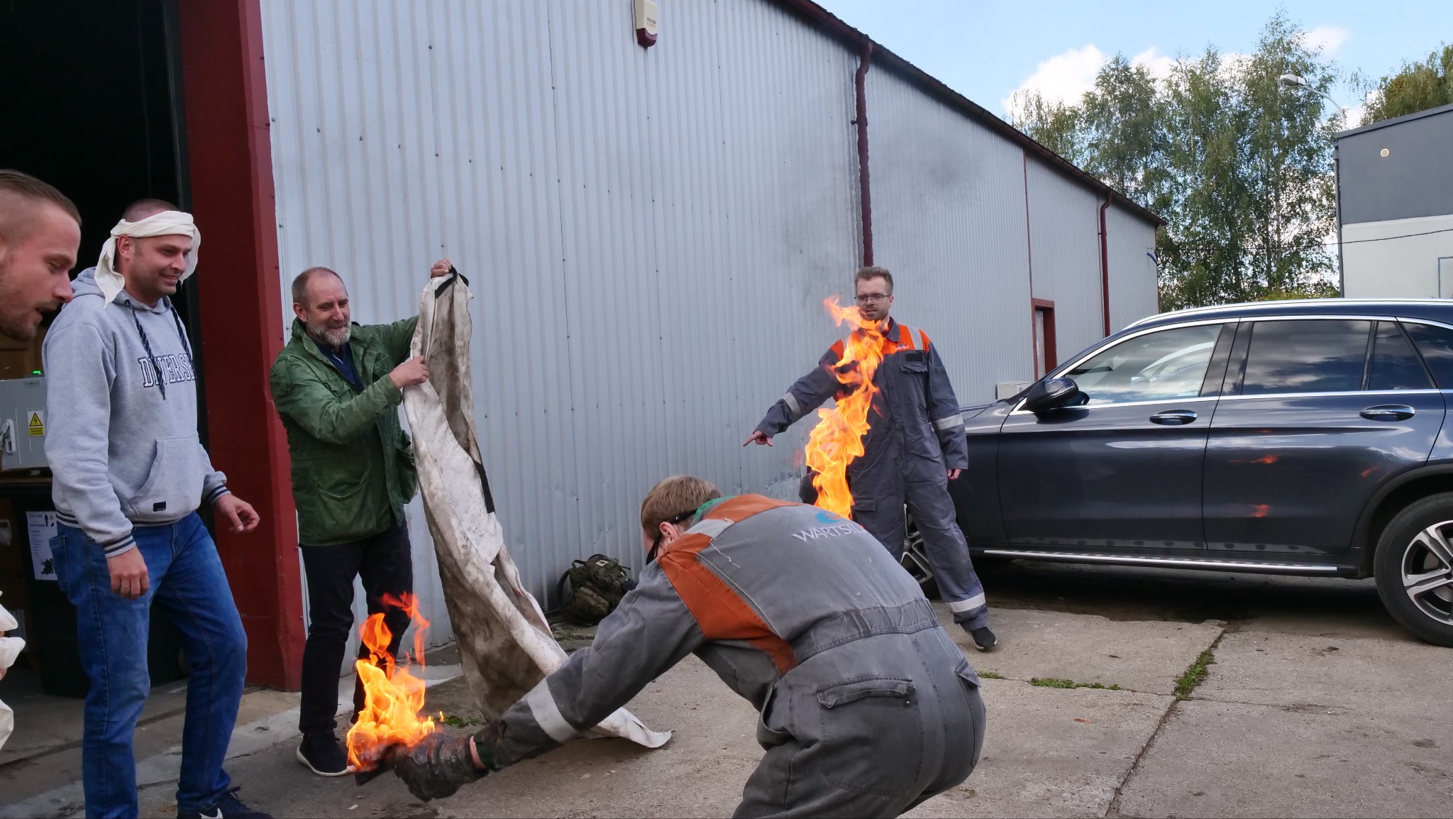 Instruktor z płomieniami symuluje pożar na manekinie podczas demonstracji gaszenia ognia kocem gaśniczym w trakcie kursu pierwszej pomocy, obok obserwatorzy z opatrunkiem na głowie i zaparkowany...
