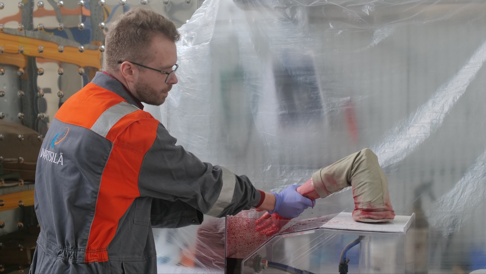 Mężczyzna w okularach i kombinezonie z logo Wärtsilä, w gumowych rękawiczkach, ćwiczy tamowanie krwotoku na sztucznej kończynie w przezroczystej komorze.
