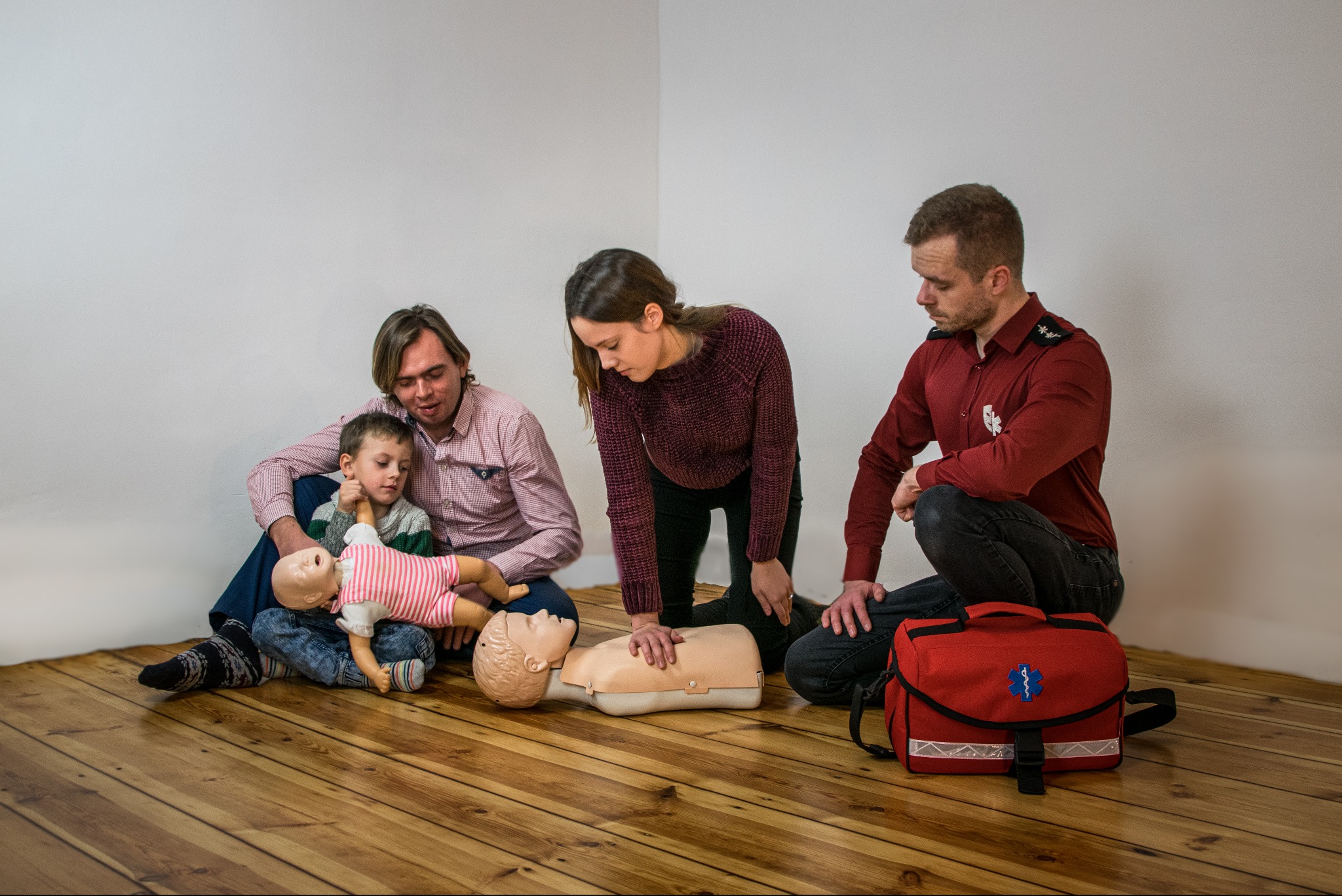 Instruktorzy demonstrujący resuscytację krążeniowo-oddechową na manekinach dla dorosłych i dzieci, z torbą medyczną w tle.