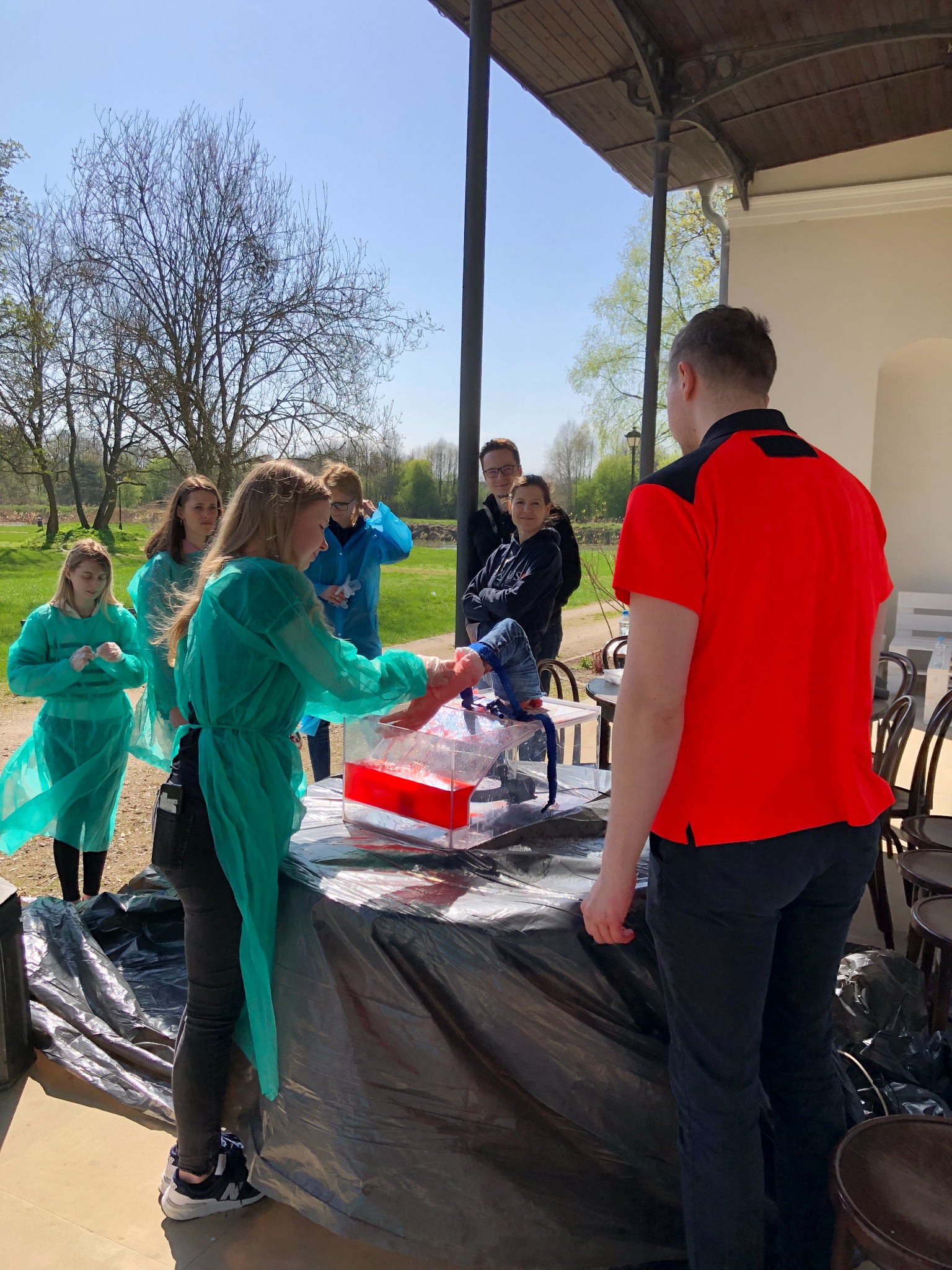 Grupa osób w zielonych fartuchach ćwiczy na fantomie umieszczonym w przezroczystym pojemniku wypełnionym czerwoną cieczą, na tle drzew i jasnego nieba.