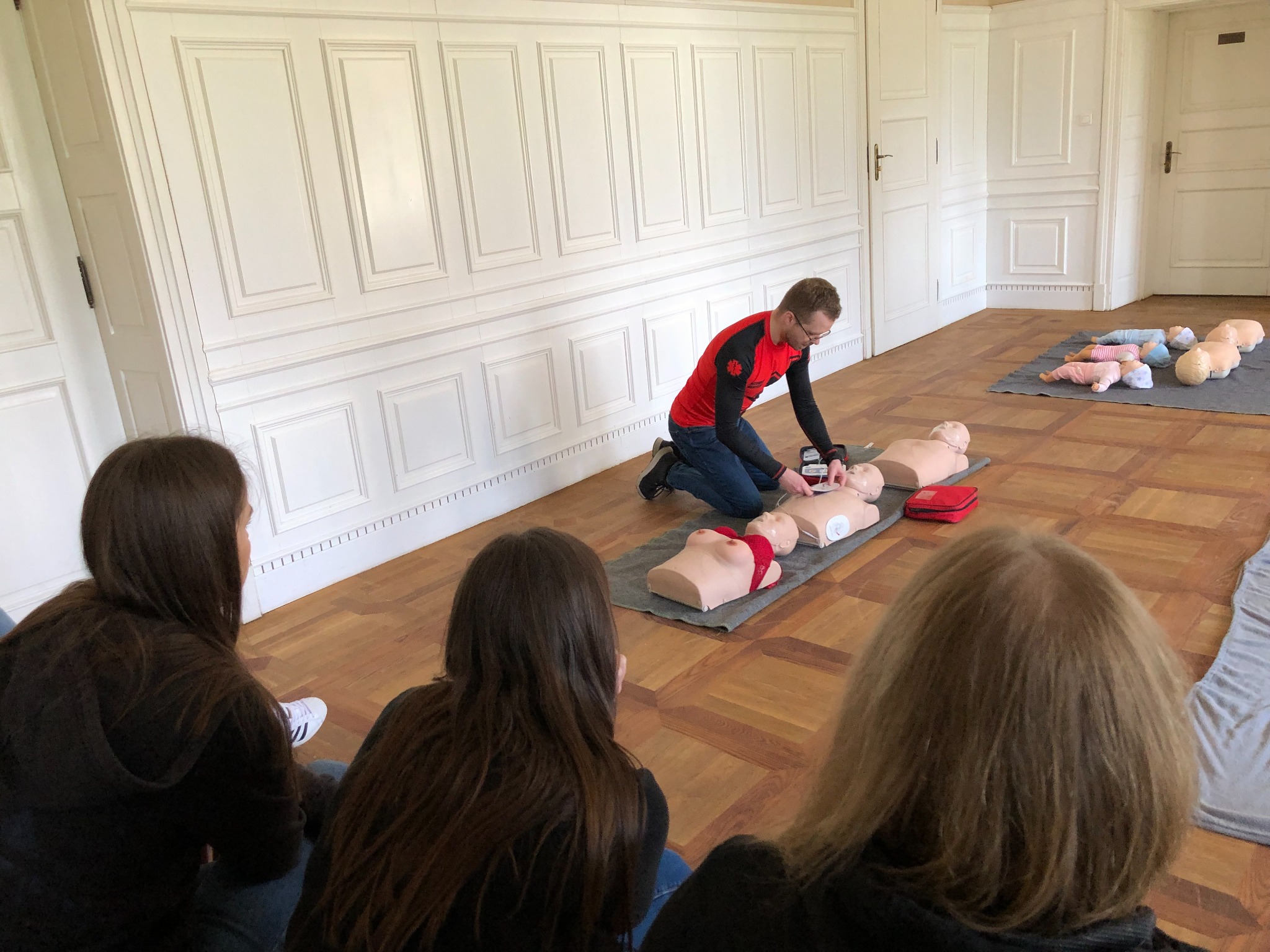 Instruktor w czerwonej koszulce demonstruje resuscytację krążeniowo-oddechową na manekinie przed grupą kursantów w przestronnym, jasnym pomieszczeniu z parkietem i białymi panelami ściennymi.